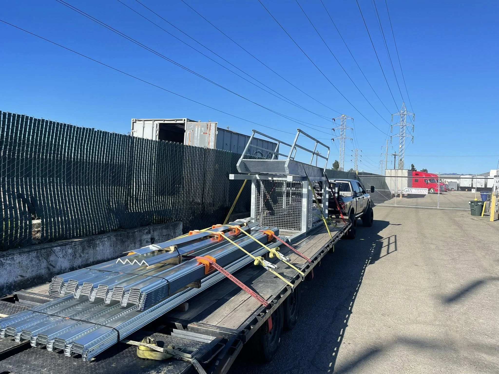 Construction trailer with metal supplies and equipment, chain-link fence, street, and electric power lines under a clear blue sky.