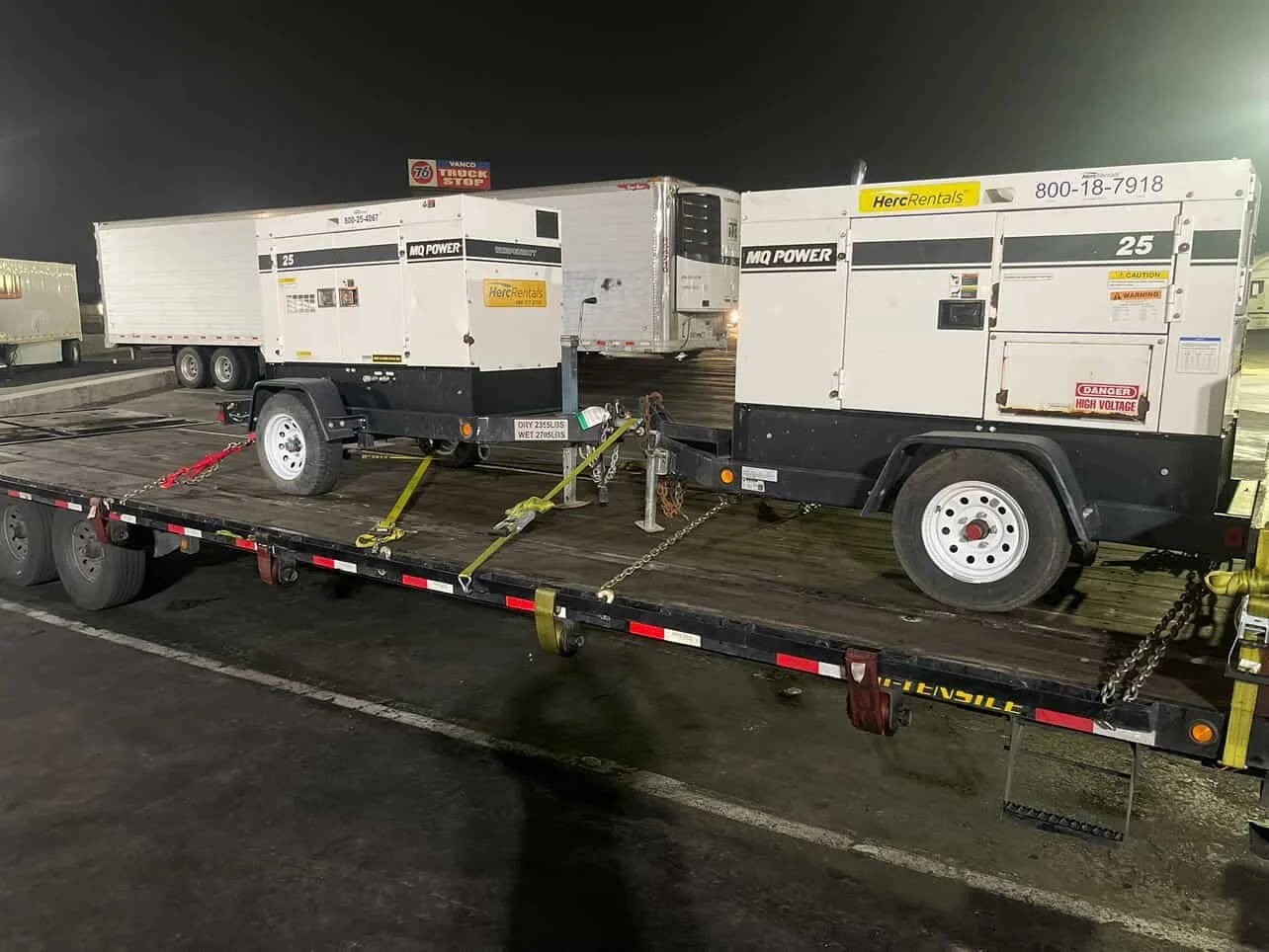 Two large mobile generators mounted on a flatbed trailer parked in a lot at night, secured with straps and chains, with other trailers and trucks visible in the background.