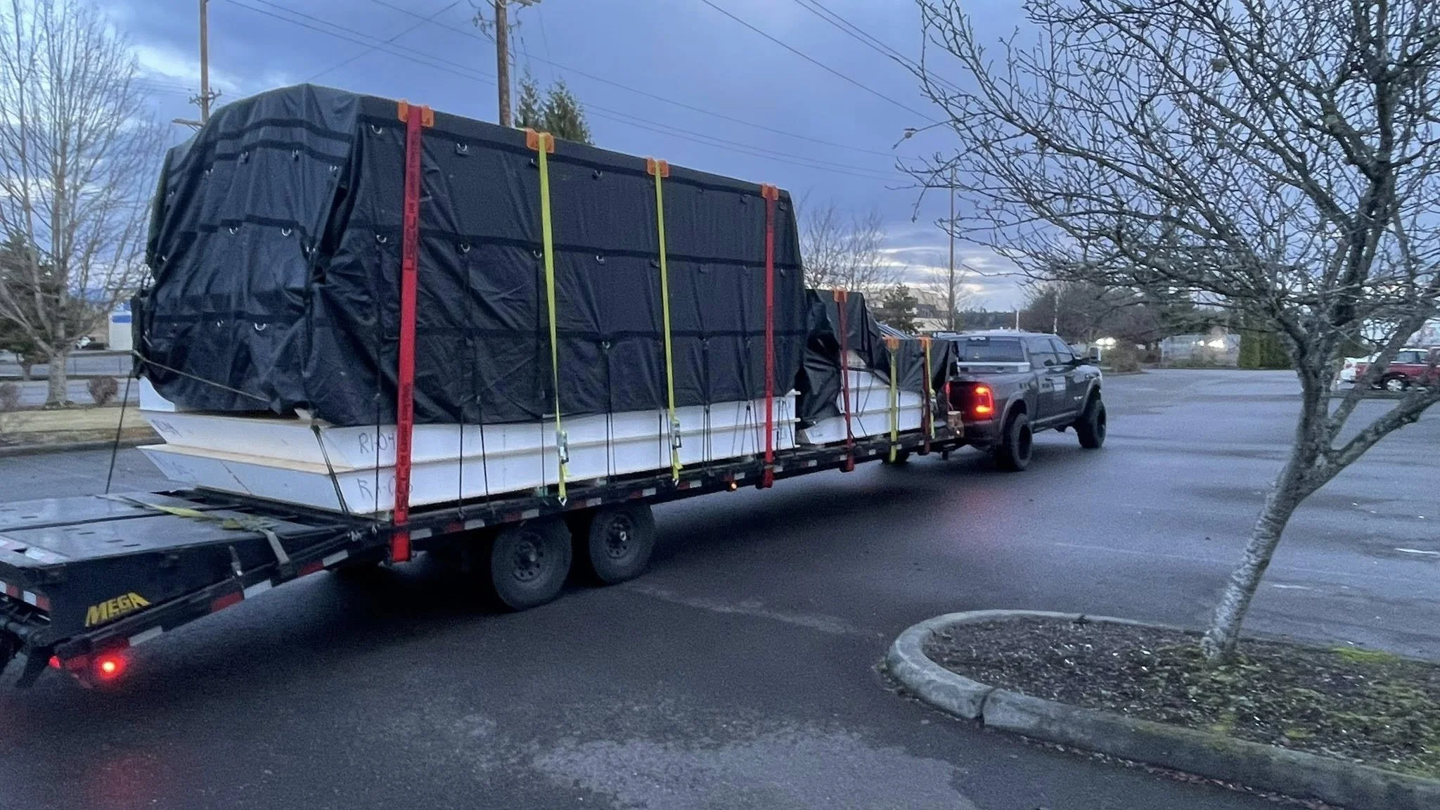 A flatbed truck carrying several large objects covered with black tarps, secured with colorful straps, parked in a parking lot near trees and other vehicles.