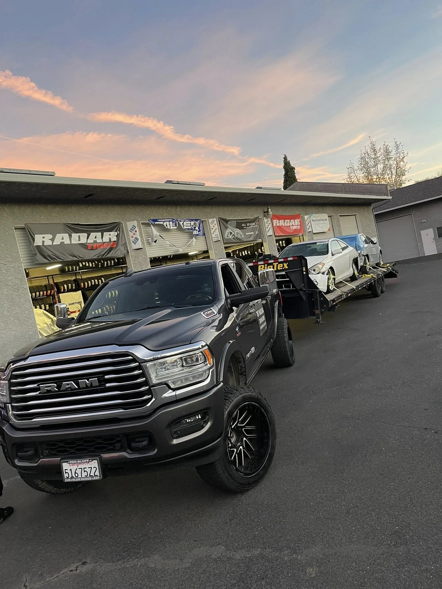 A black RAM pickup truck parked in front of an auto repair shop, with a flatbed trailer carrying two white cars, all against a sunset sky with pink and blue clouds.