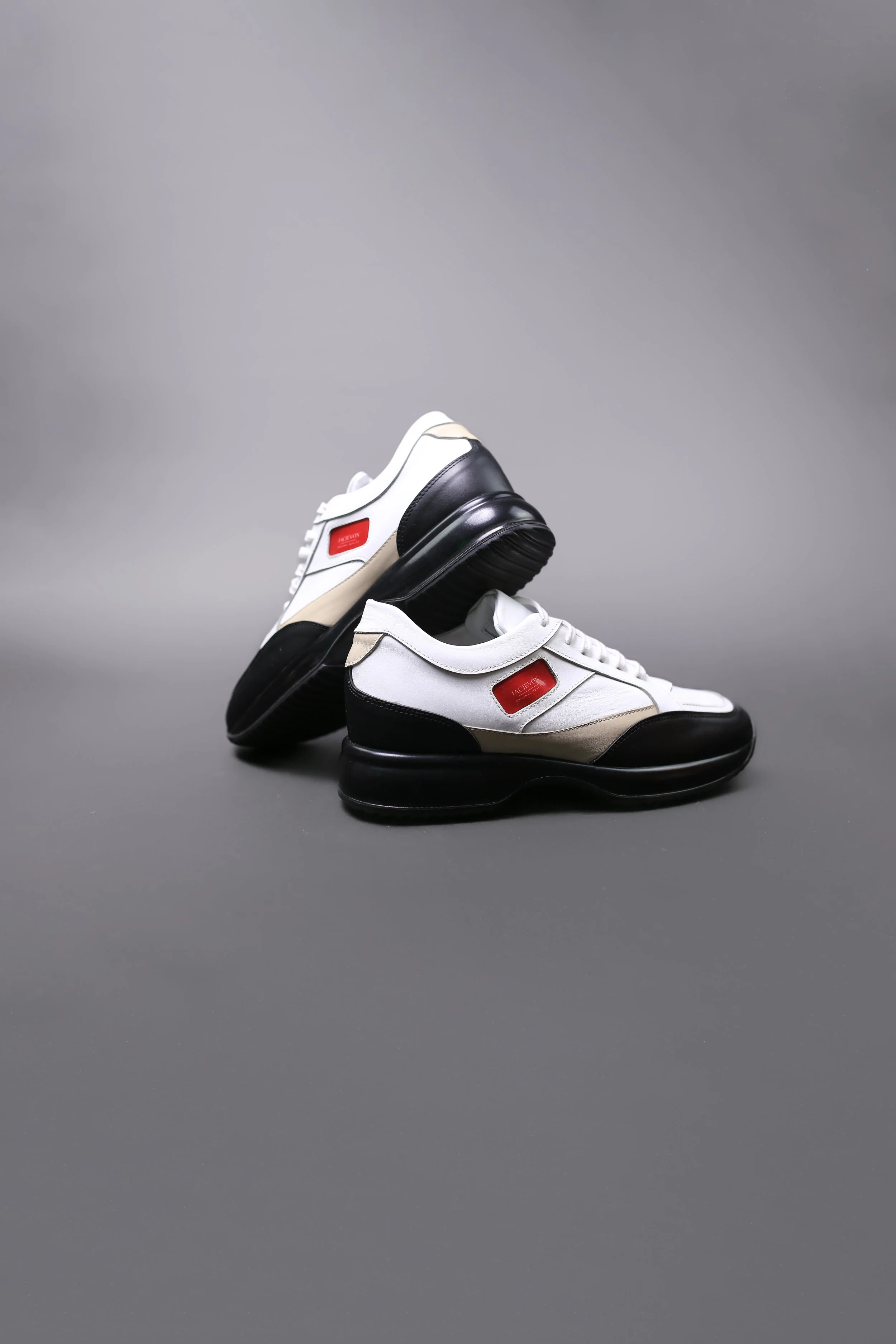 A pair of white, black, and beige sneakers with red accents, positioned on a gray surface with one shoe resting on top of the other.