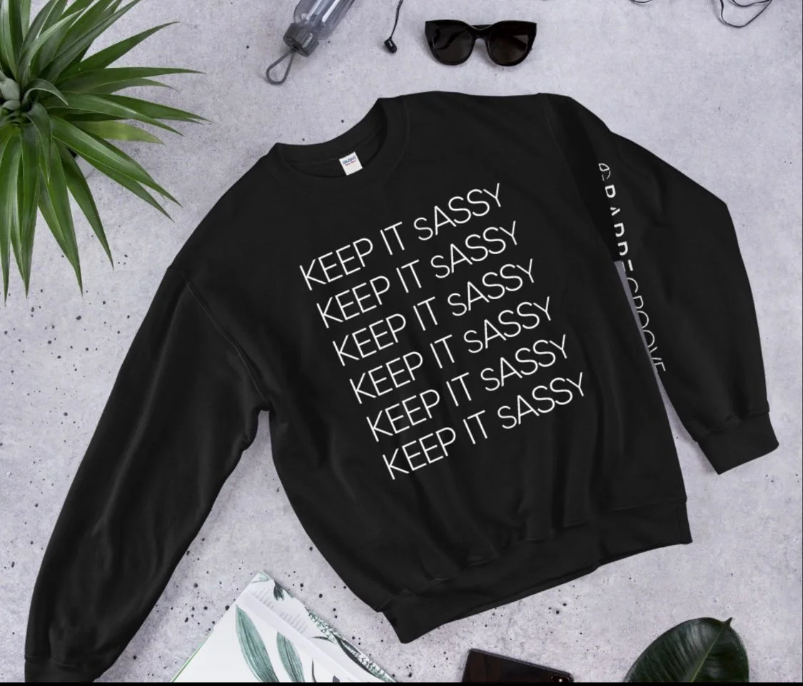 Black sweatshirt with white text reading 'Keep It Sassy' repeated multiple times, sunglasses, a potted plant, a magazine with green leaf illustrations, and a brown notebook on a gray concrete surface.