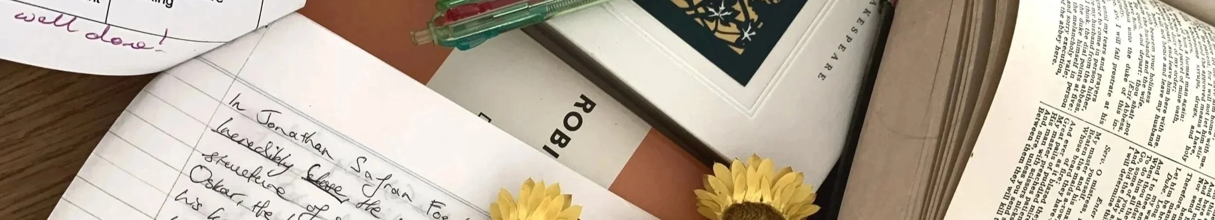 A cluttered desk with handwritten notes, a closed book, a pen, and yellow flowers placed on top of the notes. The essay feedback reads "Well done!"