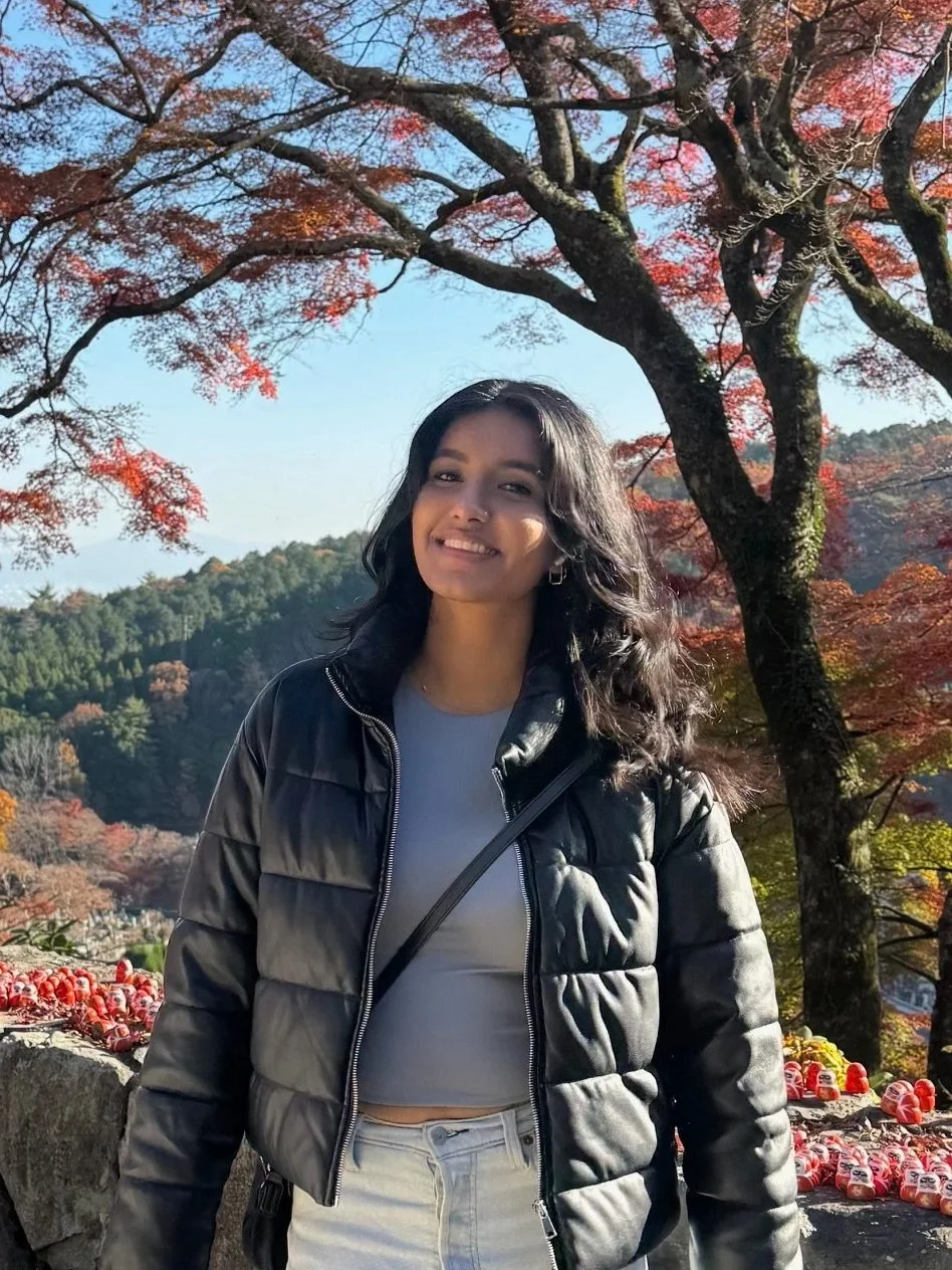 A young smiling woman wearing a black puffer jacket stands in front of a view of beautiful autumn foliage