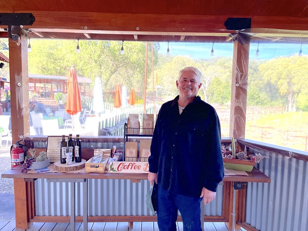 A smiling man standing in front of a wooden and metal table with coffee bags and bottles, in an outdoor setting with trees and patio umbrellas in the background.