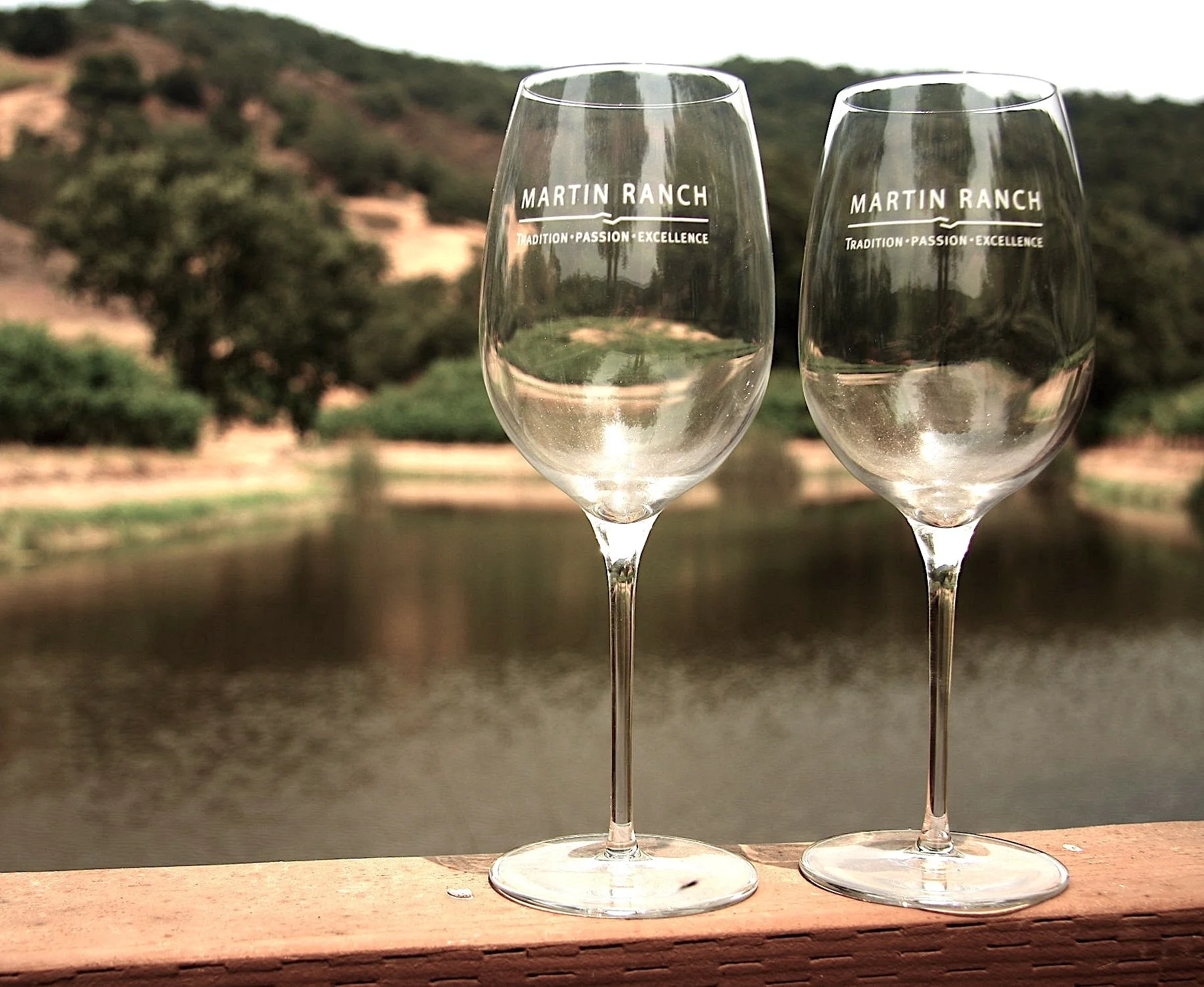 Two empty wine glasses with 'Martin Ranch' printed on them, set on a wooden surface outdoors with a blurred background of trees and hills.