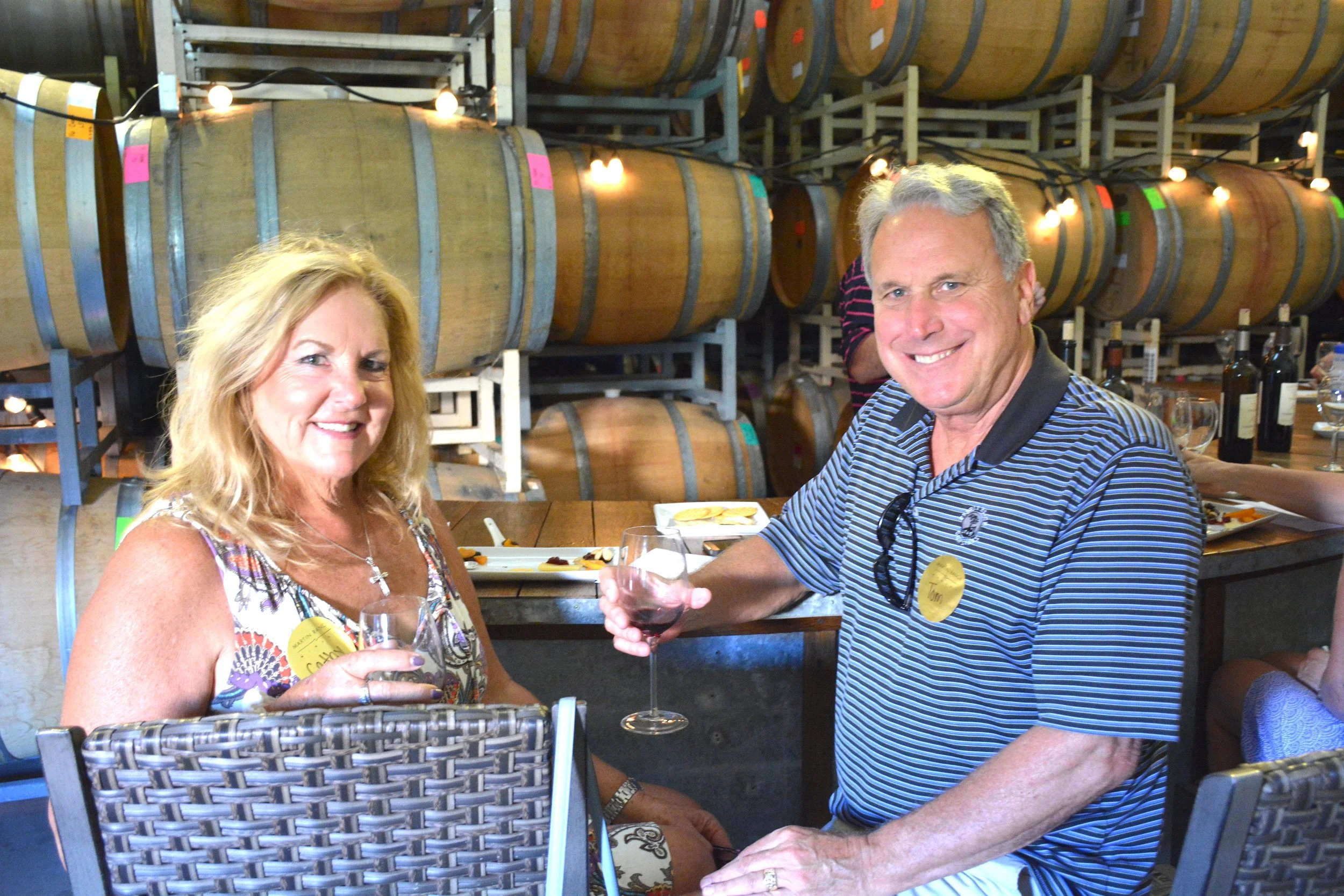 A man and woman at a wine tasting event, smiling and holding glasses of red wine, with wine barrels and bottles in the background.