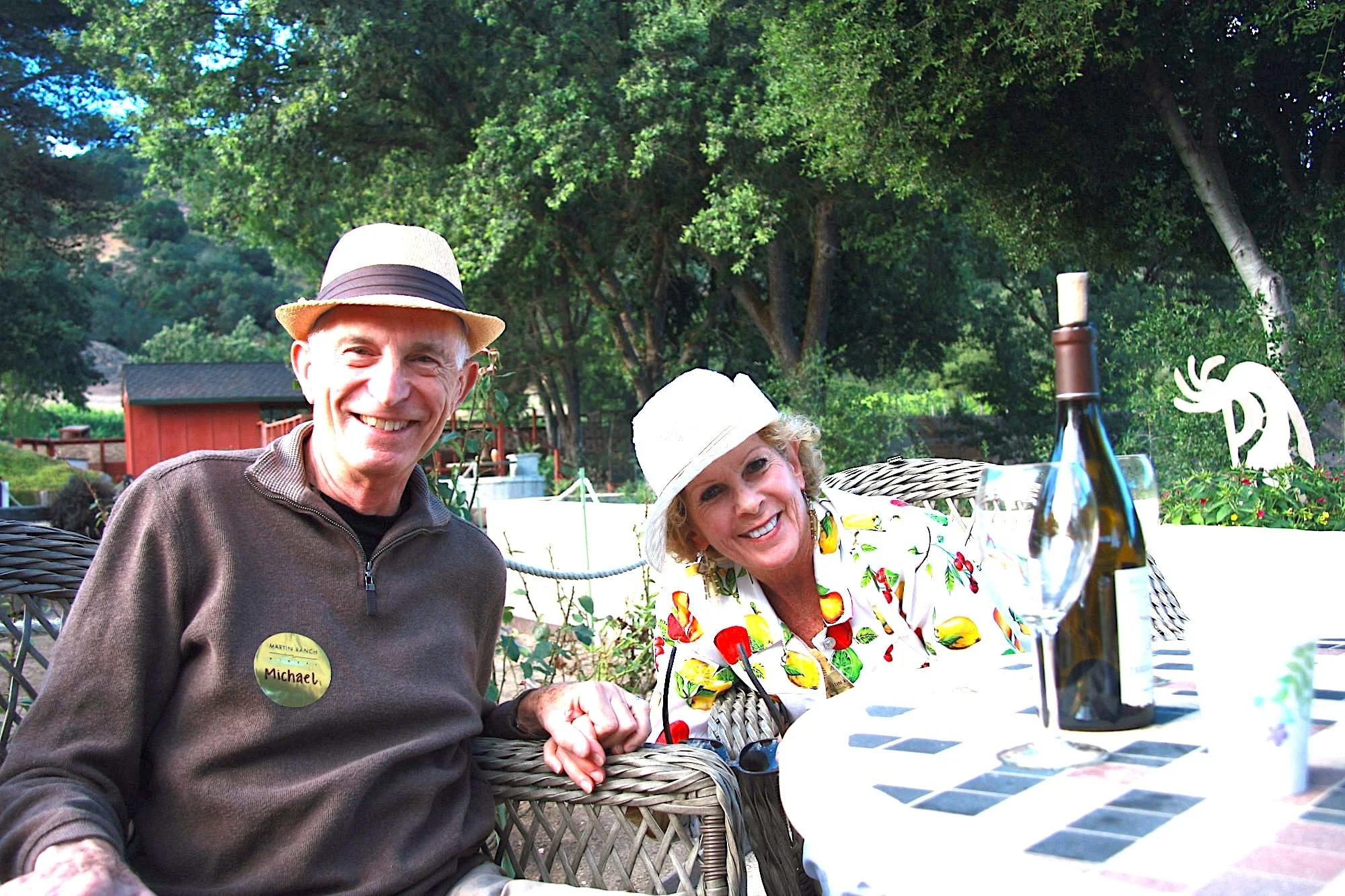 A smiling elderly man with a name tag that says "Michael" and a woman in a white hat with a yellow and red floral shirt sitting outdoors at a table with a black and white checkered tablecloth, with trees and a red building in the background.