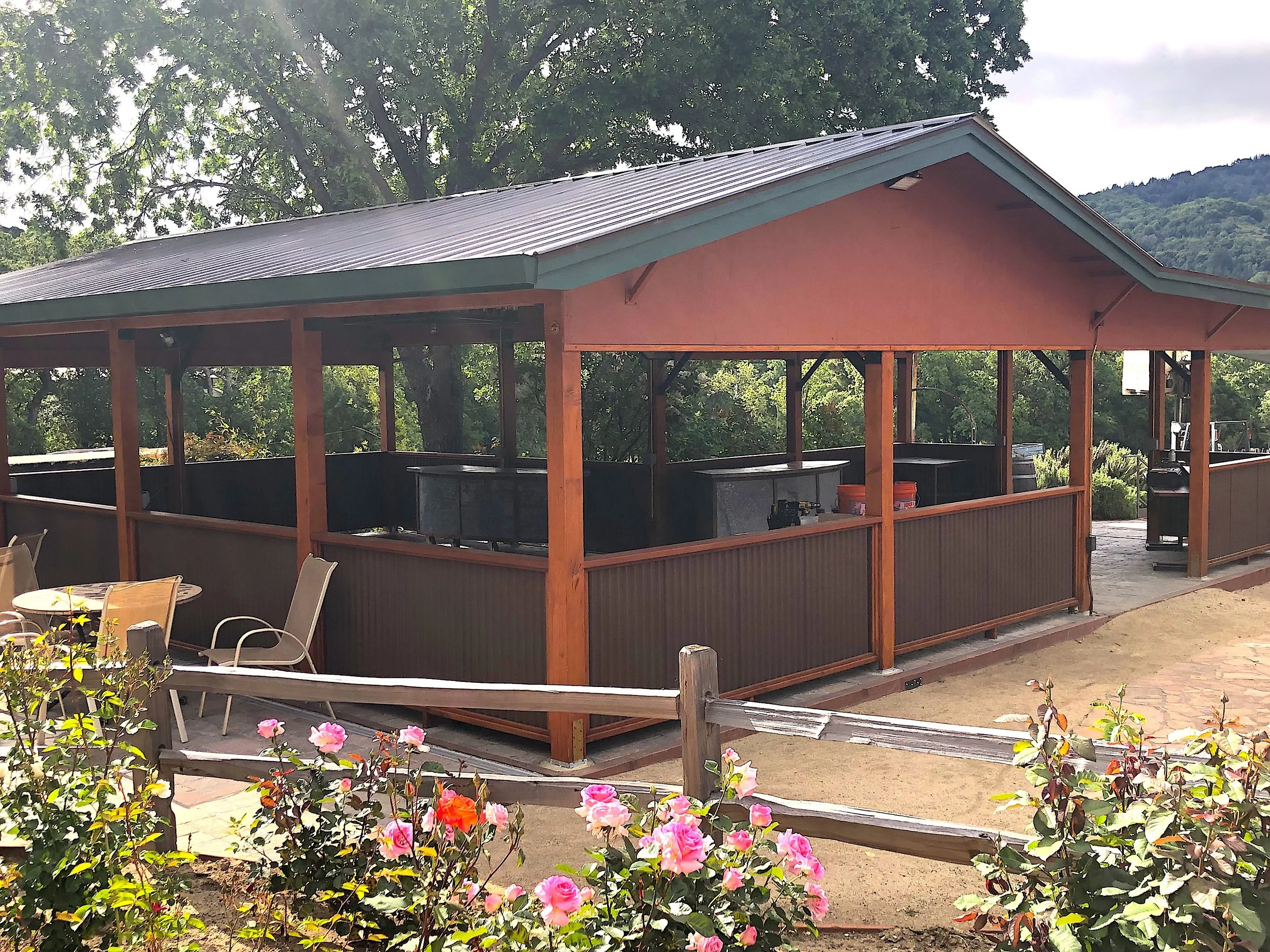 A wooden outdoor bar structure with a metal roof, set against a backdrop of lush green trees and mountains. The area features seating including chairs and tables, with colorful pink and orange flowers in the foreground.