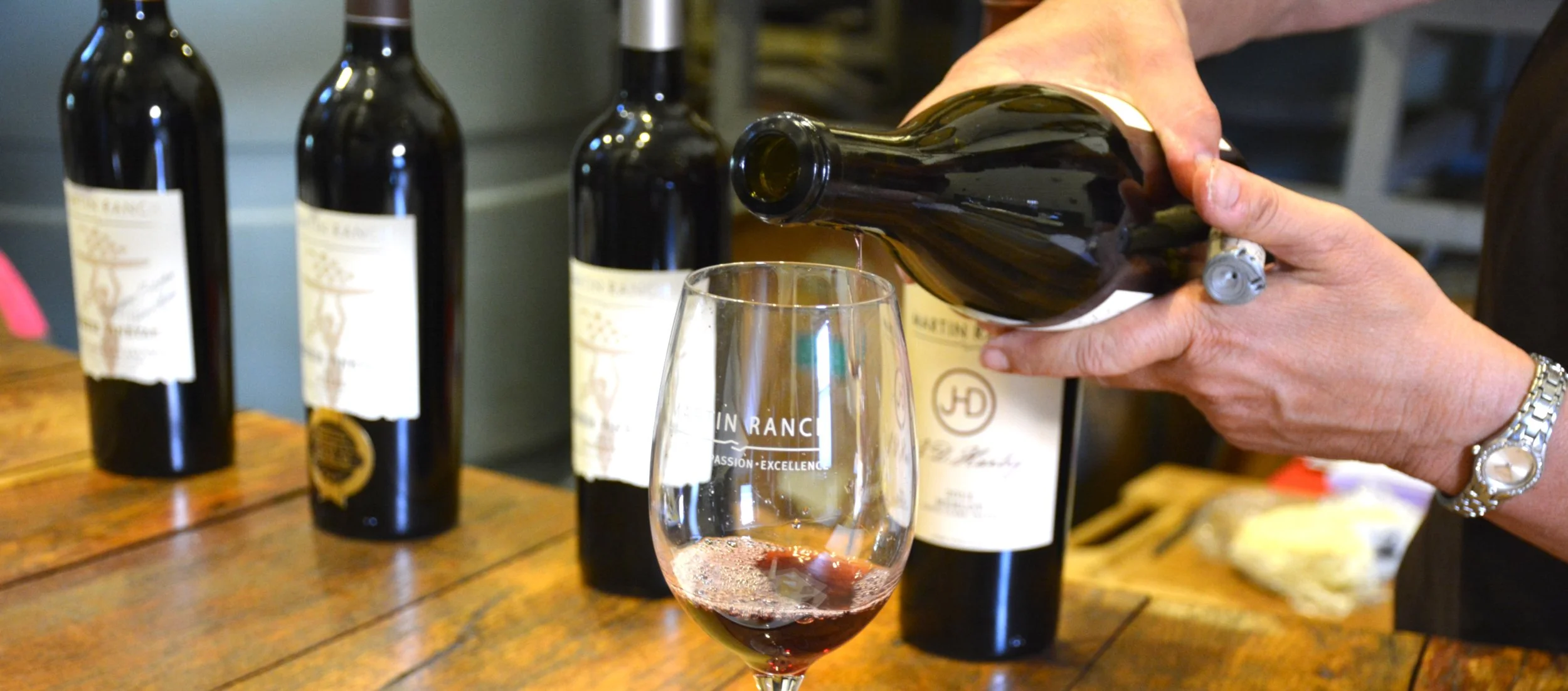 Person pouring red wine into a glass with three bottles of wine in the background on a wooden table.