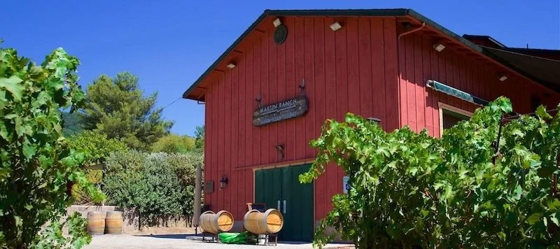 A red barn with a green door, surrounded by grapevines and barrels, under a clear blue sky.