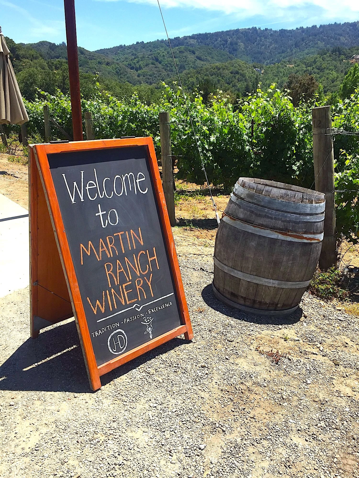 A blackboard sign reading "Welcome to Martin Ranch & Winery" with a vineyard and green wooded hills in the background, and a large wine barrel beside it.