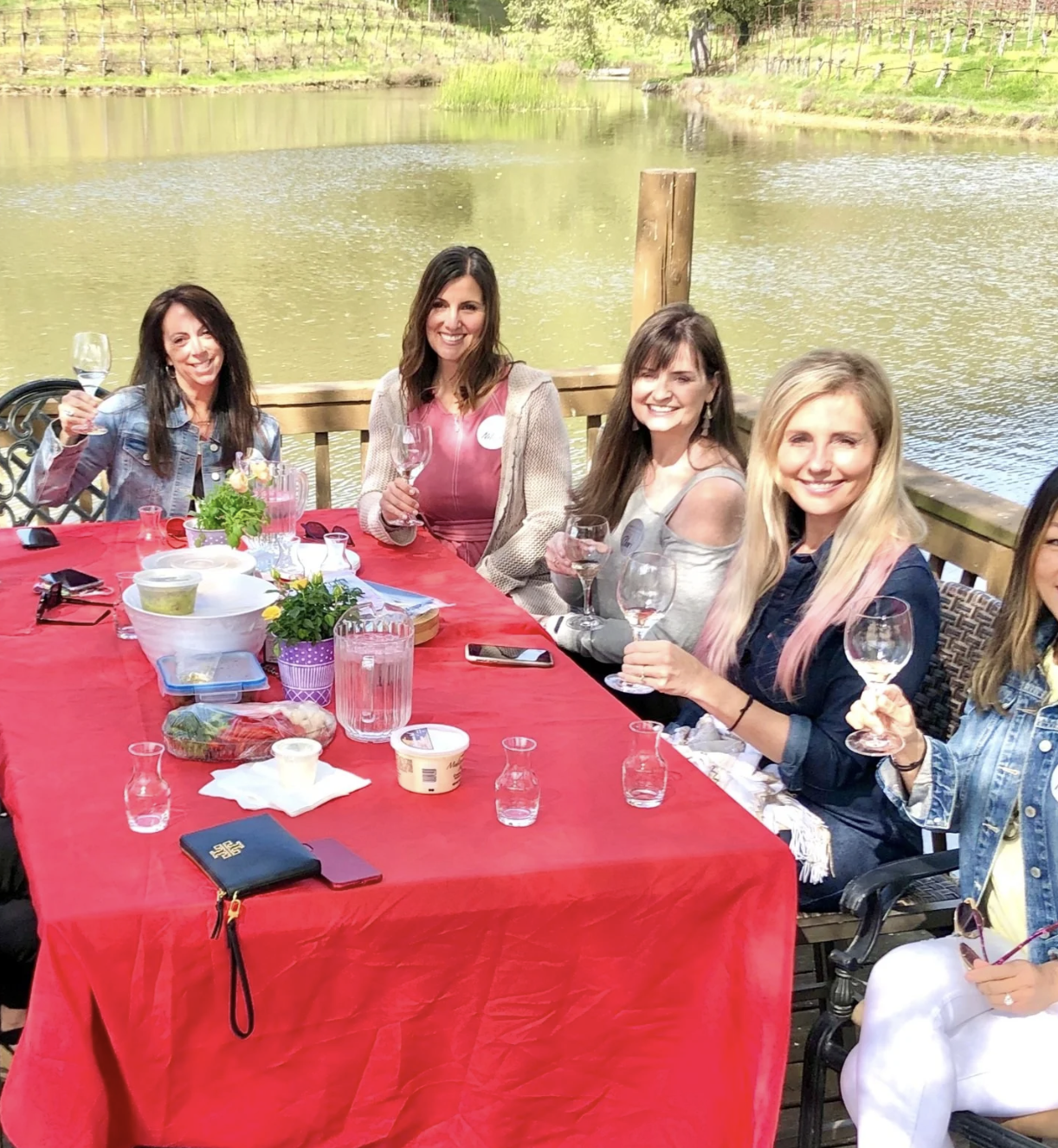 A group of women sitting around a table with a red tablecloth outdoors by a lake, holding wine glasses and smiling.