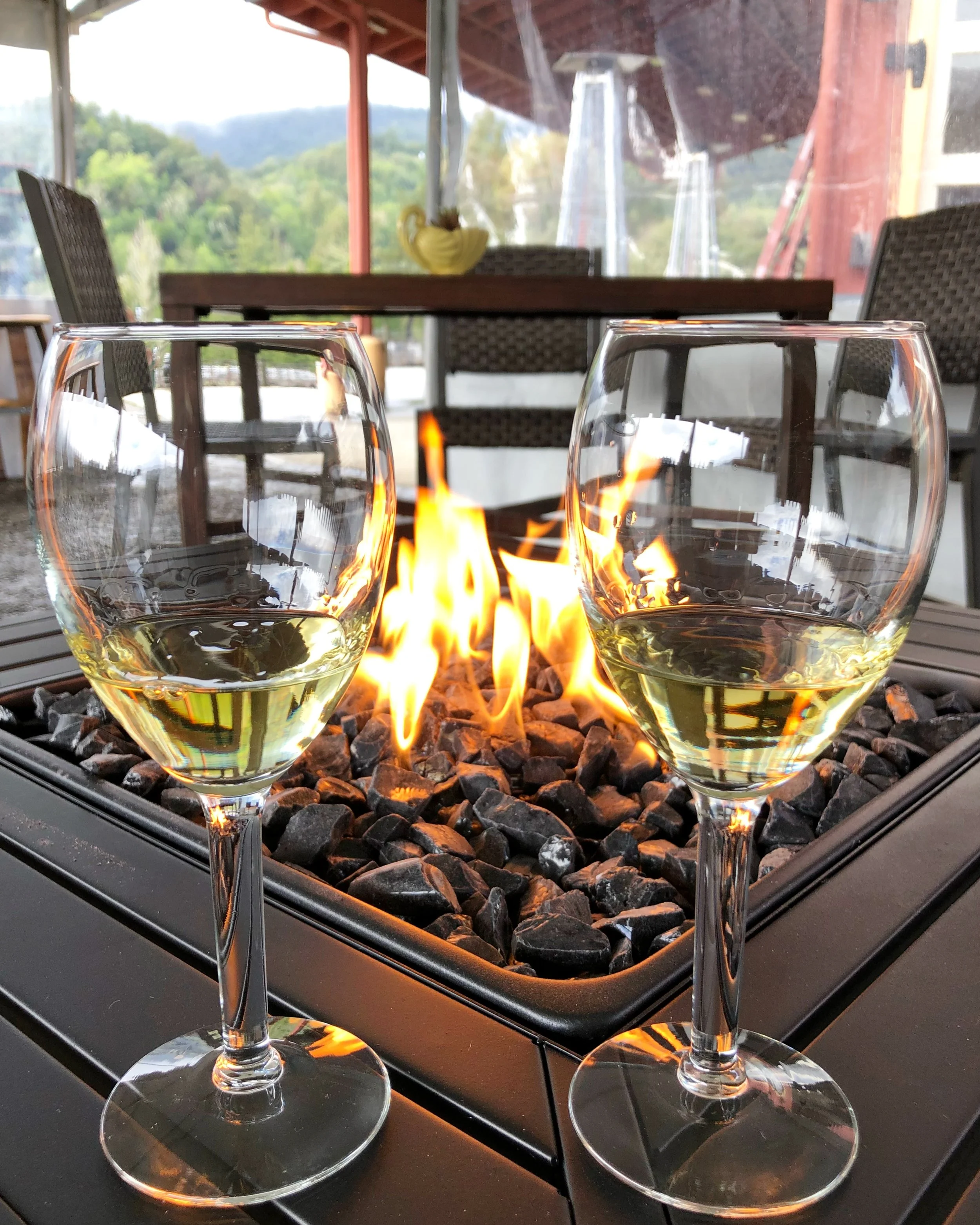 Two glasses of white wine on a table in front of a fire pit with flames and rocks, set on a patio with outdoor furniture and a mountain view in the background.