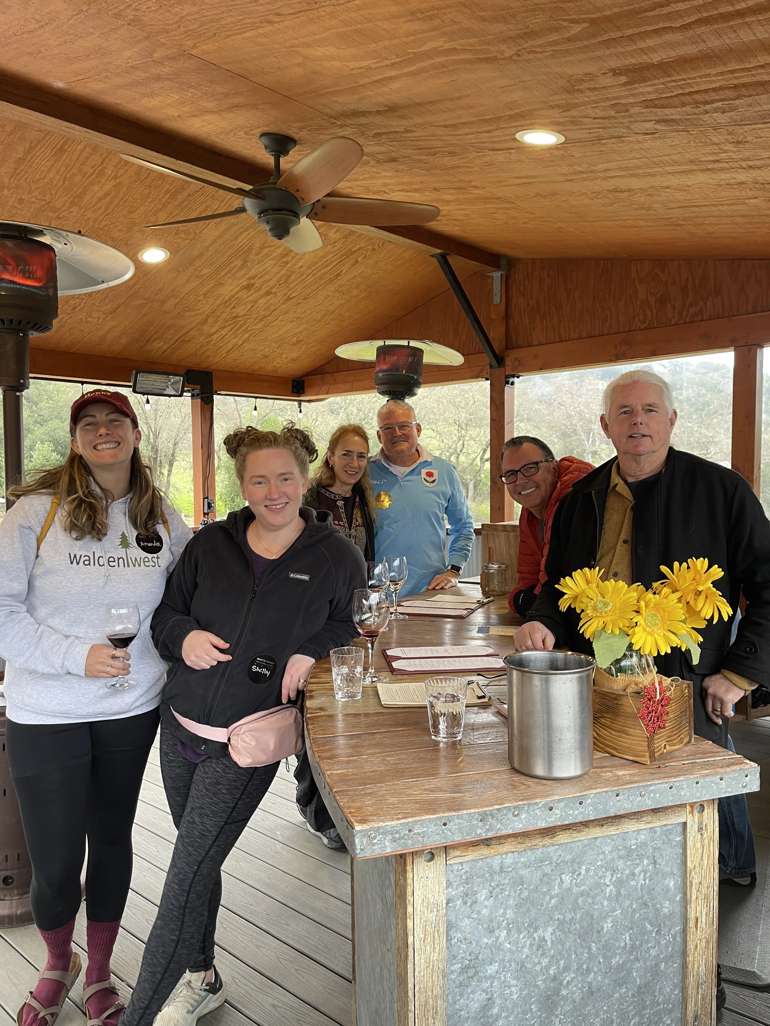 Group of six people standing around a wooden table with yellow flowers, wine glasses, and menus, inside a wooden pavilion with ceiling fans and heaters, outdoors beyond the pavilion.