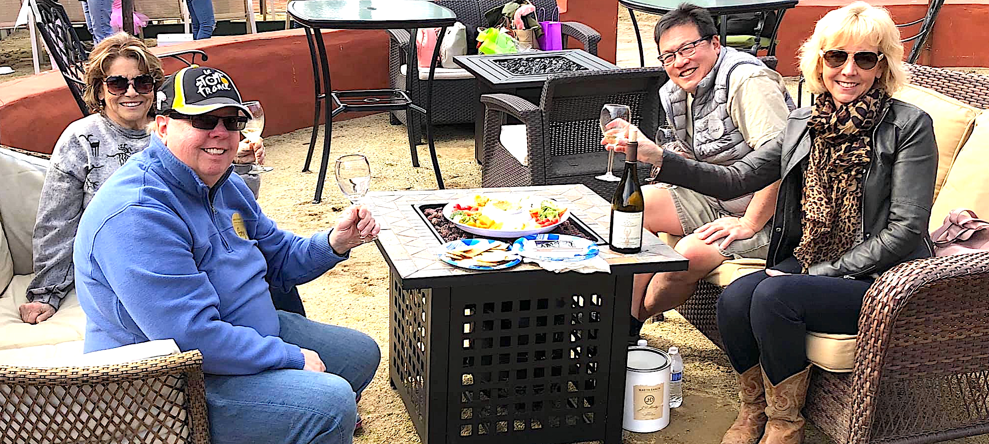 A group of five people sitting outdoors at a table, smiling, with drinks and a platter of food in front of them, enjoying a social gathering.