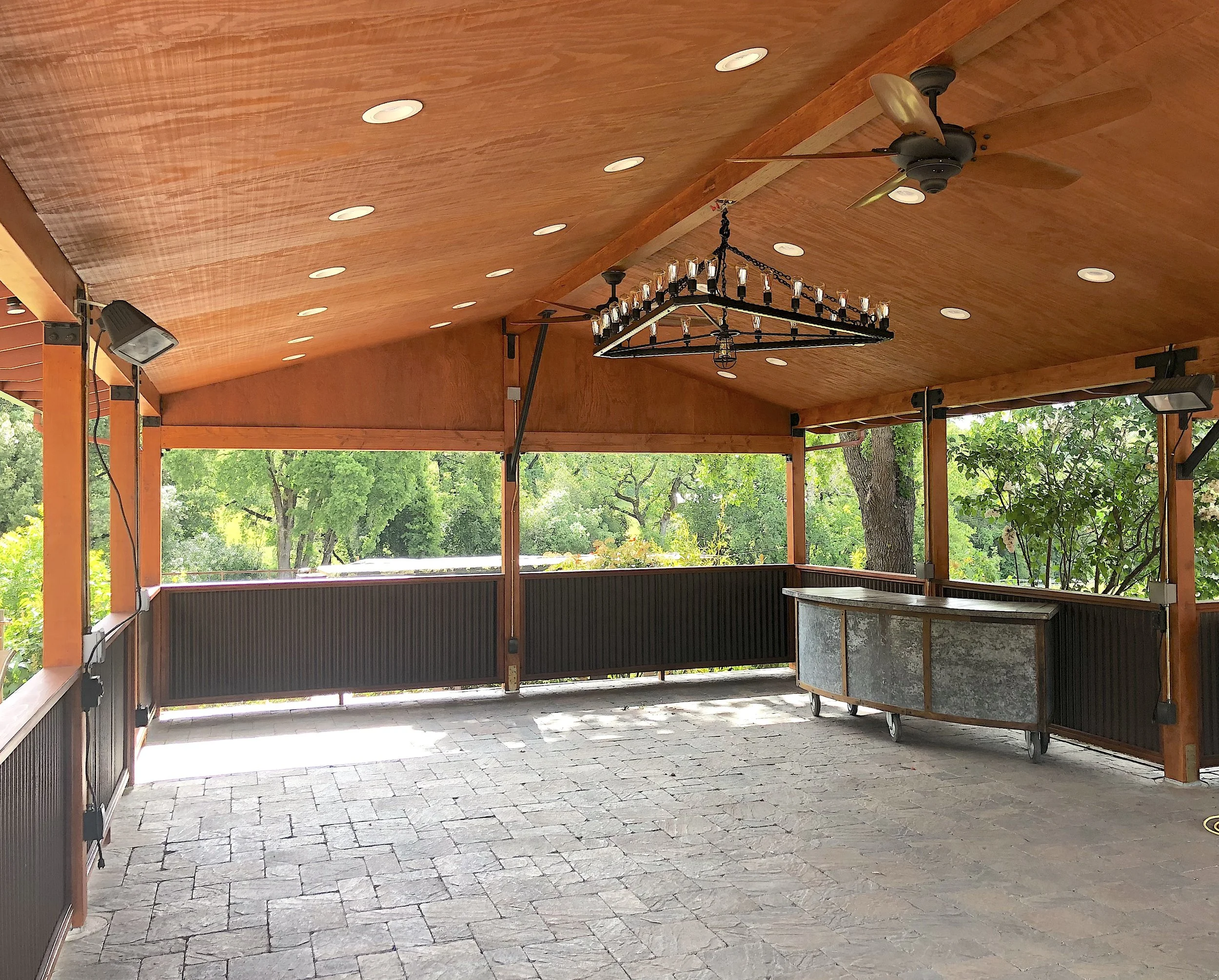 Open outdoor pavilion with a wooden ceiling, ceiling fan, chandeliers, and outdoor lighting, surrounded by green trees.
