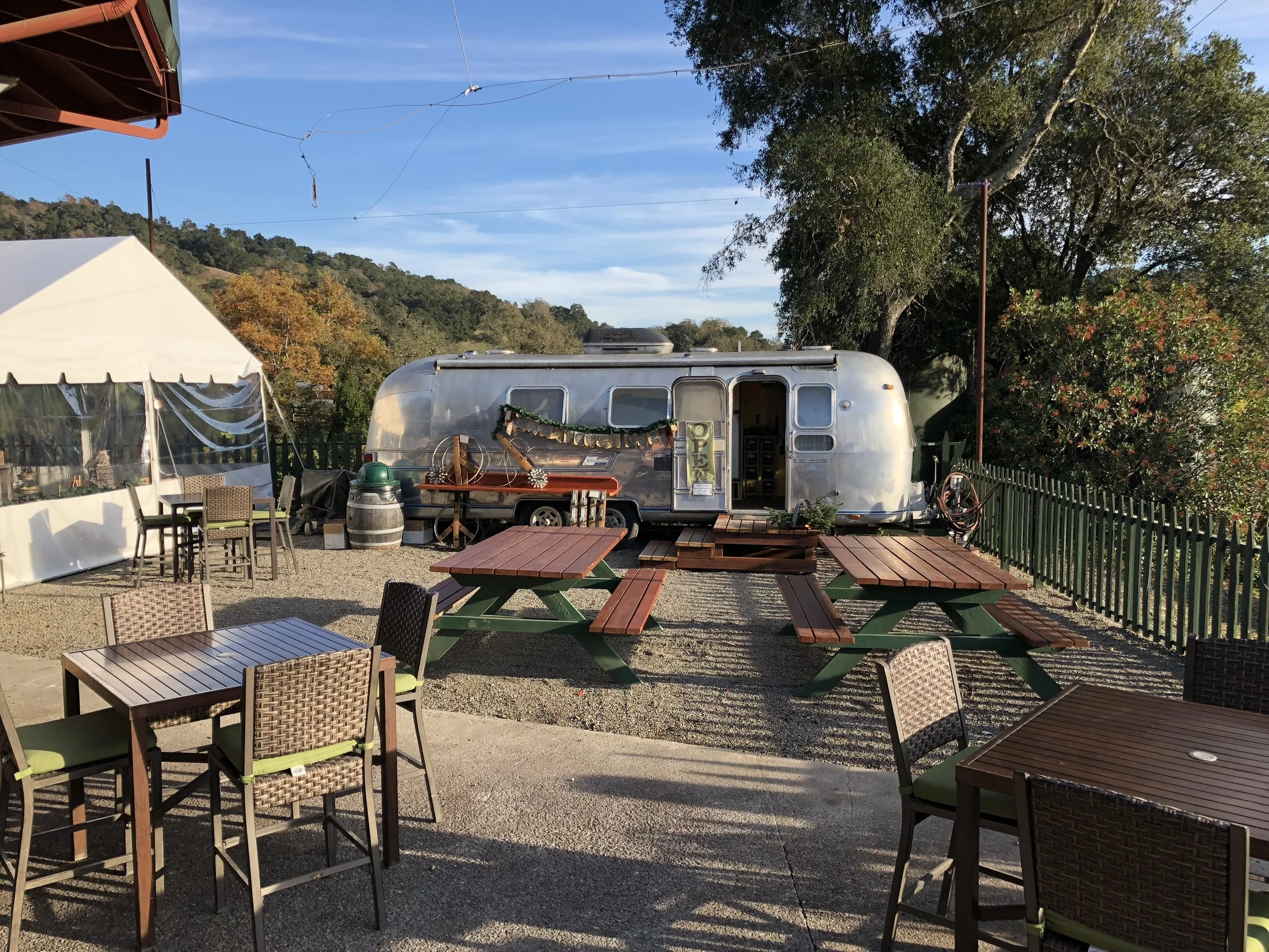 Outdoor seating area with wooden tables and chairs, a white tent, a vintage camper trailer decorated with garlands, and large trees in the background under a blue sky with some clouds.