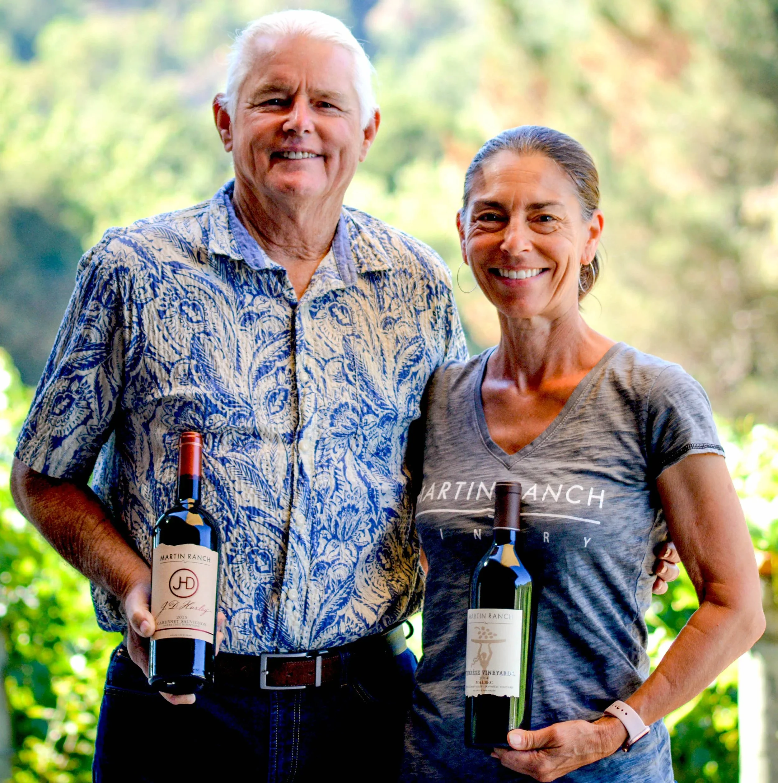 Two smiling adults, a man with white hair wearing a patterned shirt and a woman with brown hair wearing a gray T-shirt, holding bottles of wine outdoors with a green, blurred background.