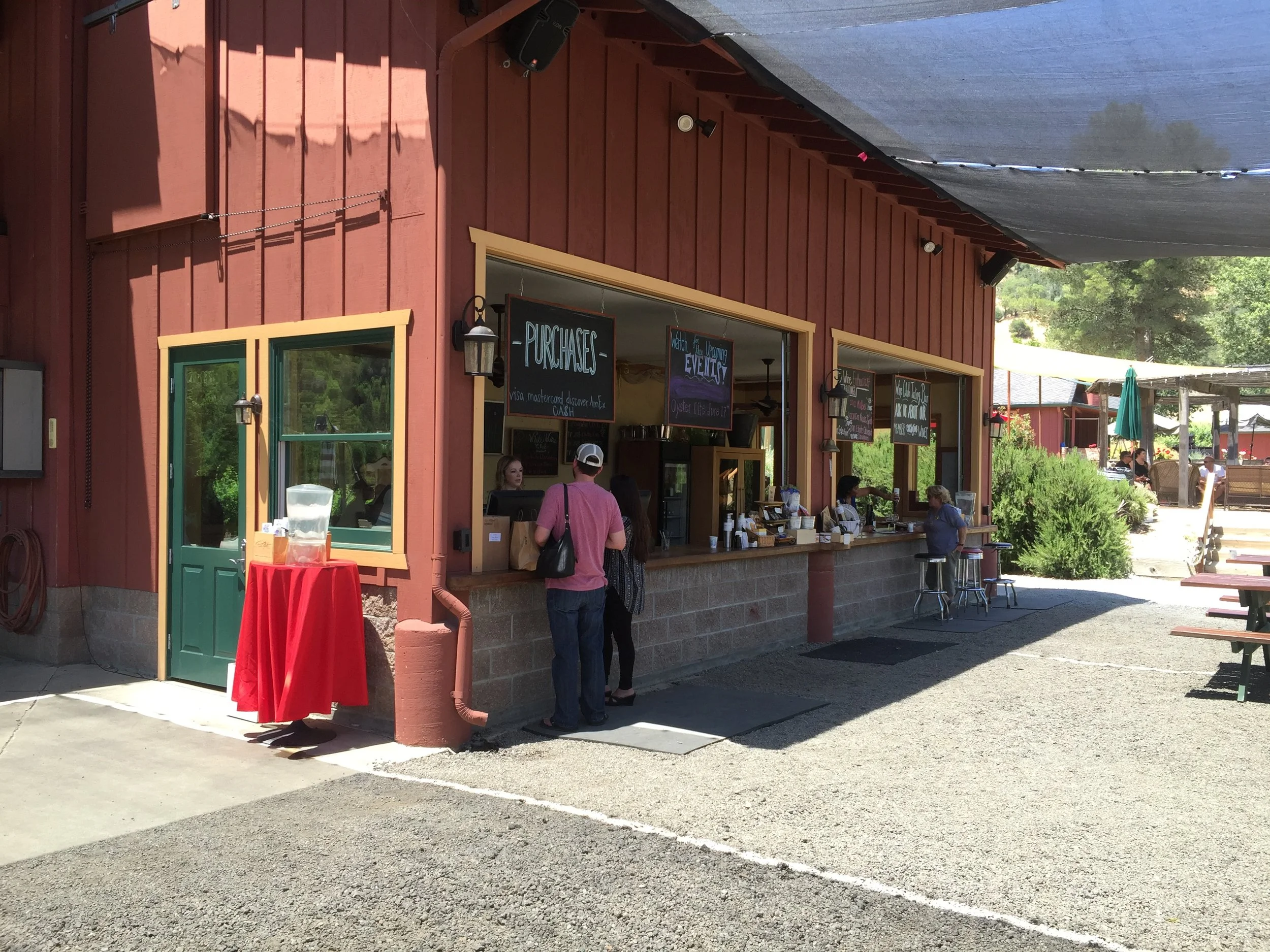 A bakery or cafe with a red wooden exterior and chalkboard menus. Customers are ordering at the counter and others are seated outside. There is a red table with a water pitcher in the foreground.