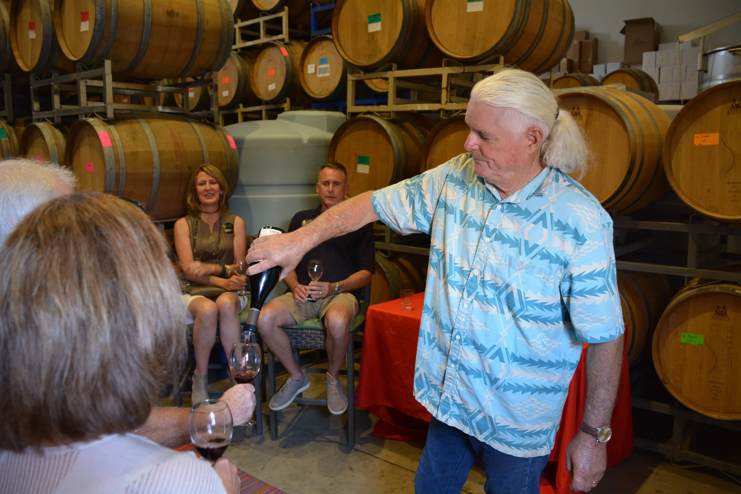 A man with long white hair in a ponytail wearing a blue patterned shirt is pouring wine into a glass for a seated woman holding a glass of red wine. Several people are sitting and standing nearby, with large wine barrels stacked on shelves in the bac