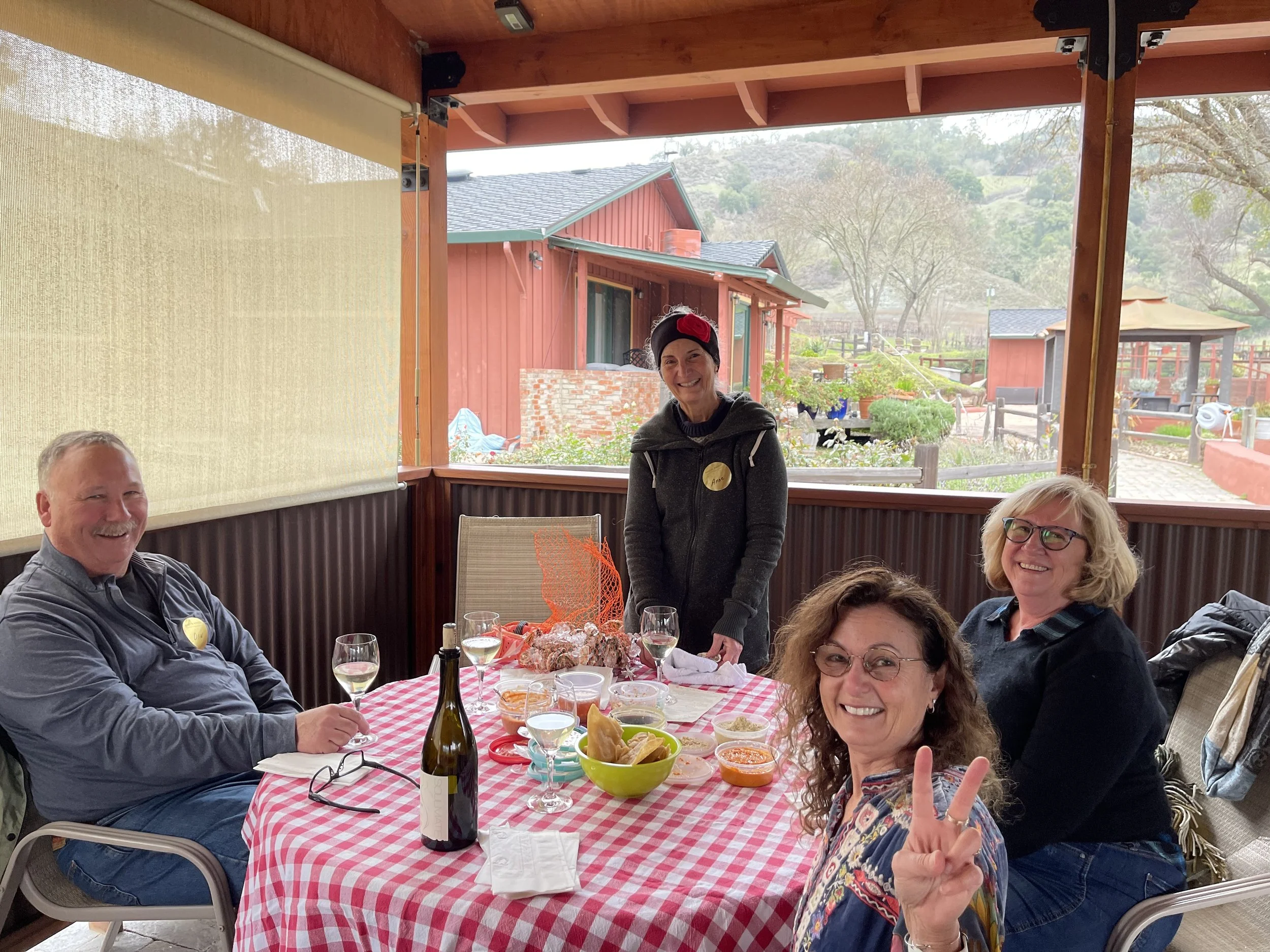 Smiling woman with curly hair wearing glasses making a peace sign at a dinner party. Other people are smiling and seated around a table with food and drinks. The scene is outdoors with a view of trees and buildings in the background.