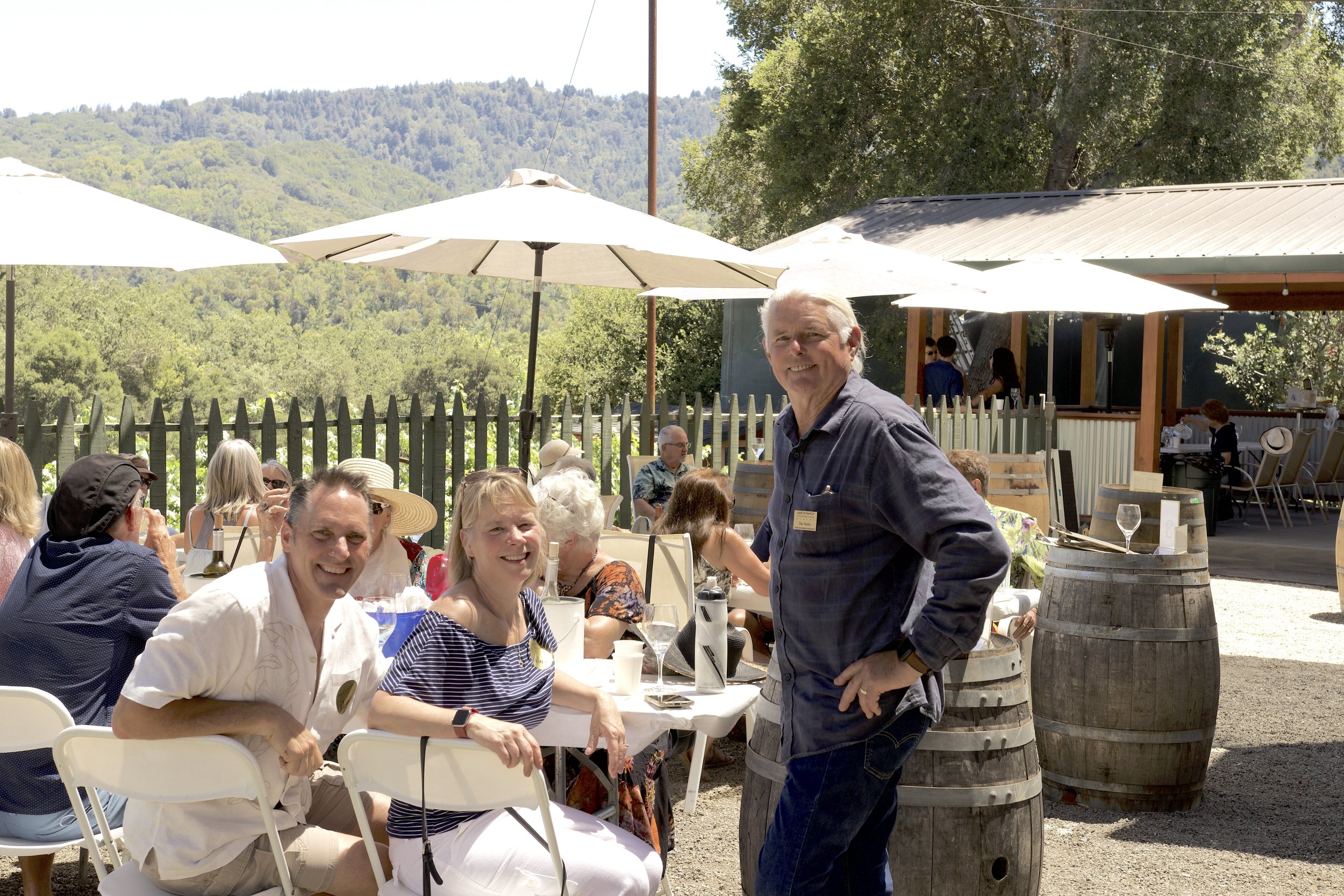 People enjoying an outdoor restaurant or winery setting with mountains and trees in the background. There are umbrellas, tables, and a man standing near a wine barrel.