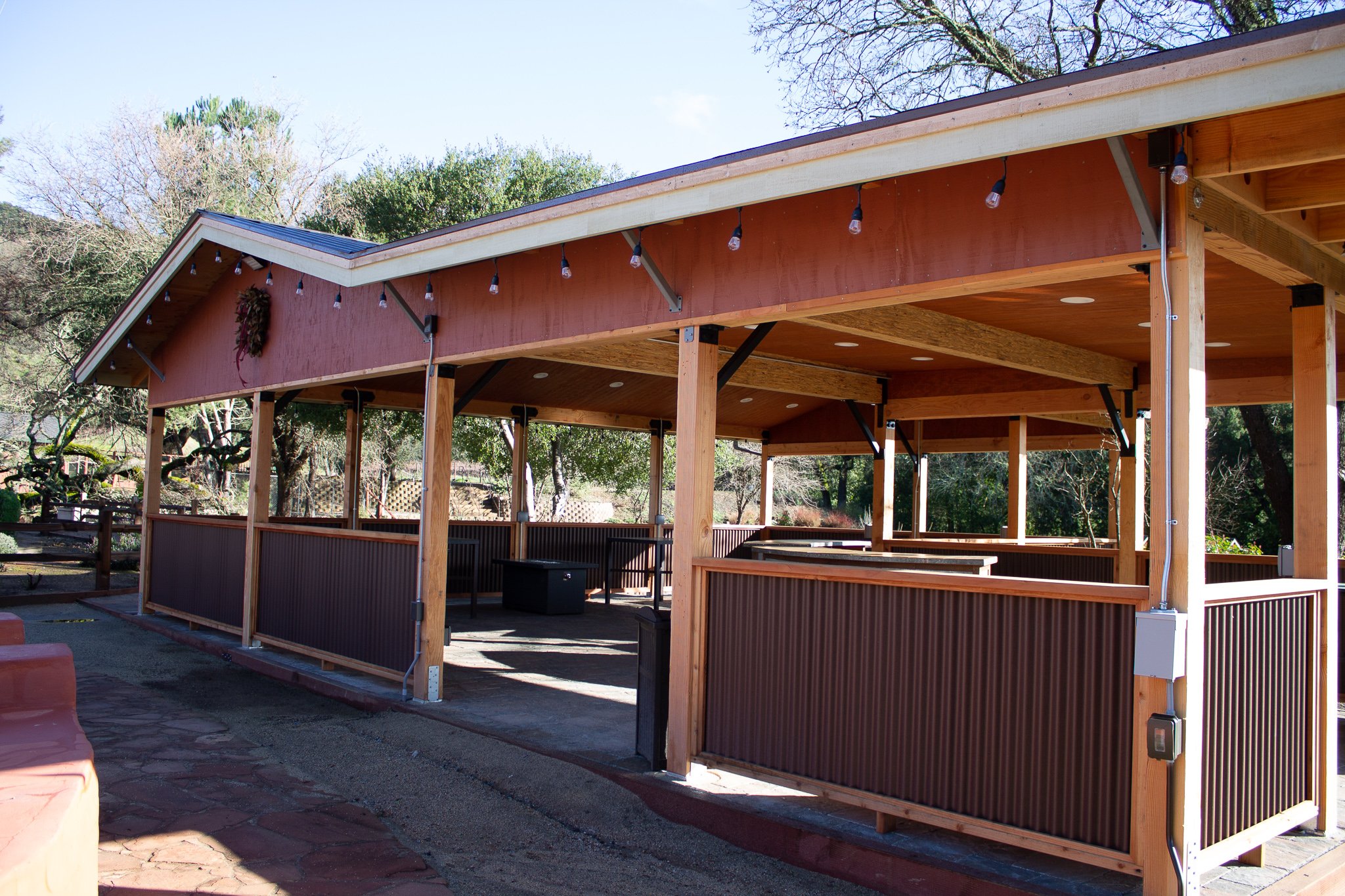 Photo of a wooden deck or pavilion with a red exterior, string lights hanging from the roof, and trees in the background.