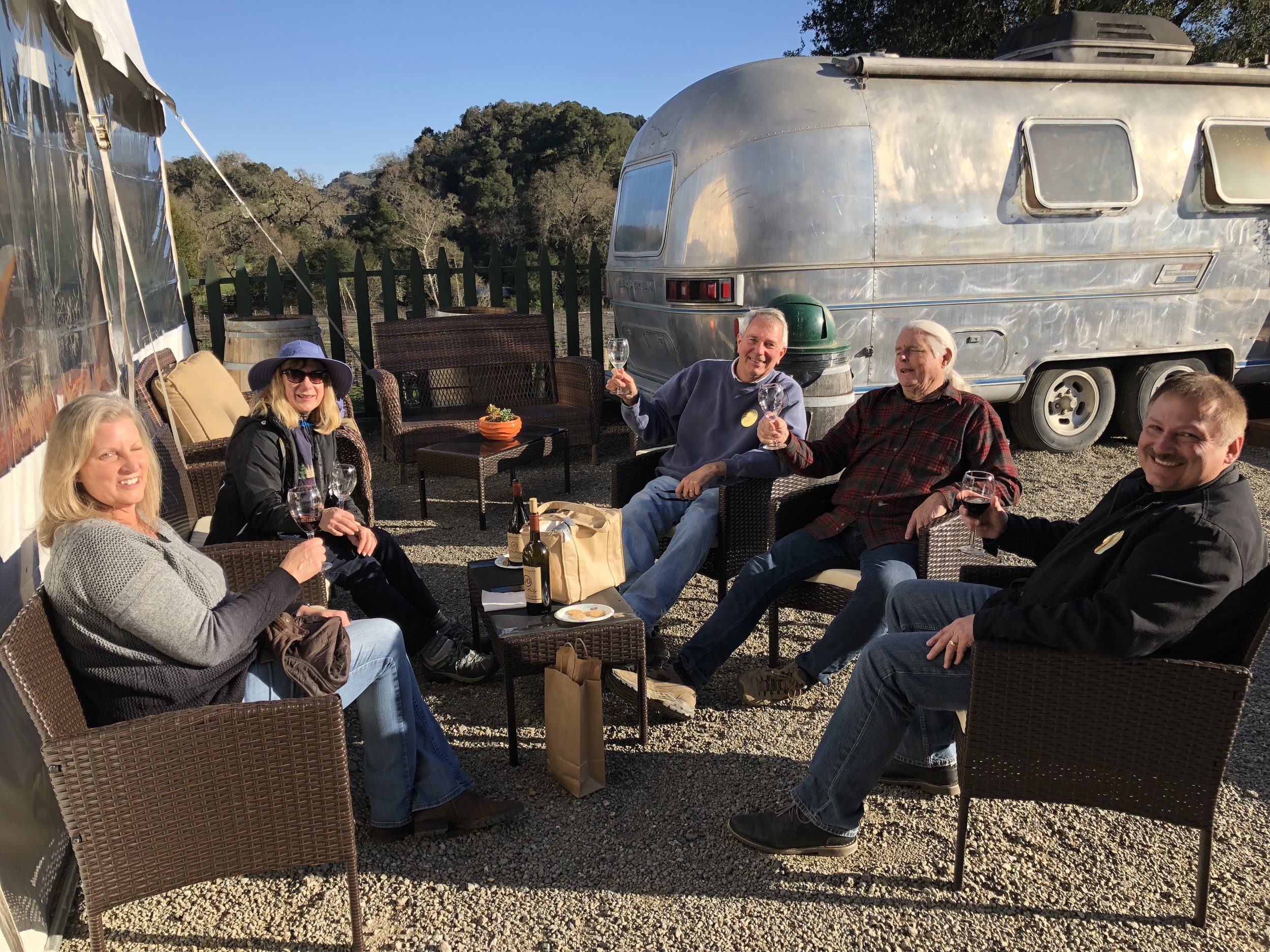 Group of people sitting outdoors at a gathering, holding wine glasses, with an RV and a tent in the background, during daytime.