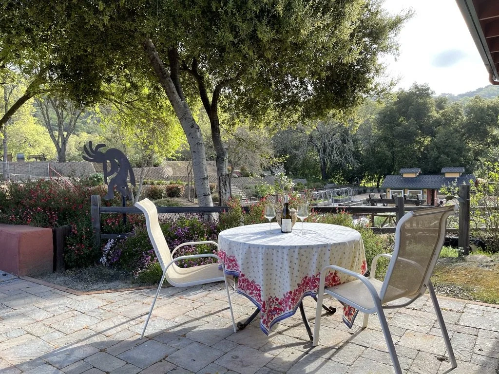 Outdoor patio scene with a round table covered with a colorful tablecloth, a bottle of wine and three glasses. Two white chairs and one beige chair surround the table. Lush green trees and shrubs in the background, with a decorative sculpture and a w