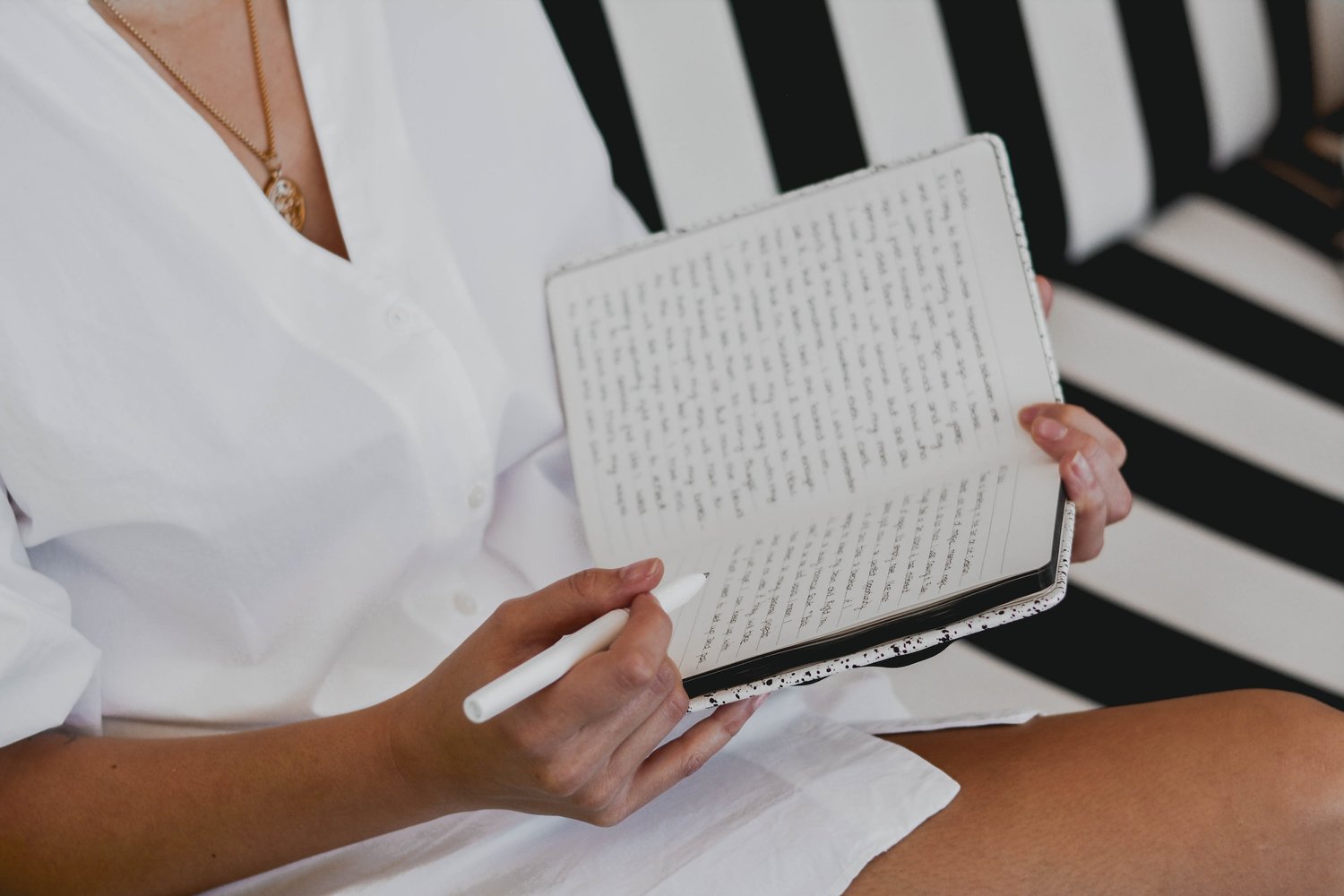 a light brown skinned person is wearing a white dress and a gold necklace. They are seated on a black and white striped couch, holding what a pens to be a journal and a pen in their right hand.