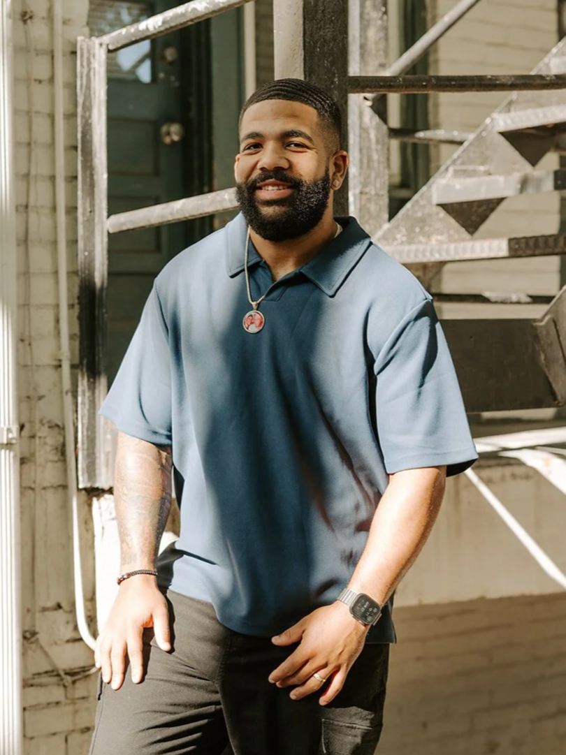 A smiling Black man with a beard, wearing a blue polo shirt, a necklace, a watch, and a bracelet, standing outdoors in front of a building with a metal staircase.