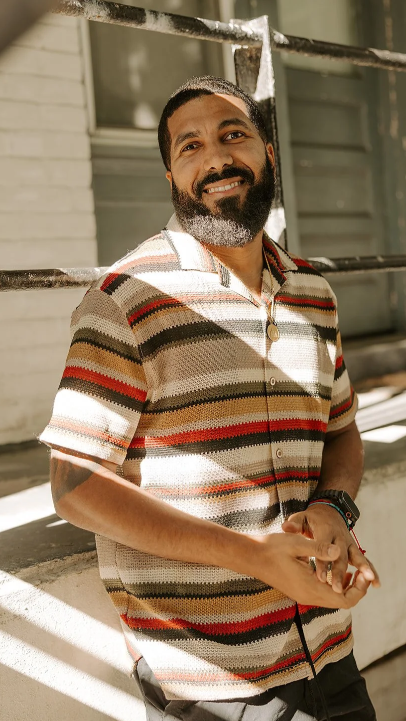 A man with a beard standing with arms crossed, wearing a black T-shirt with a bear graphic, in front of a brick wall.