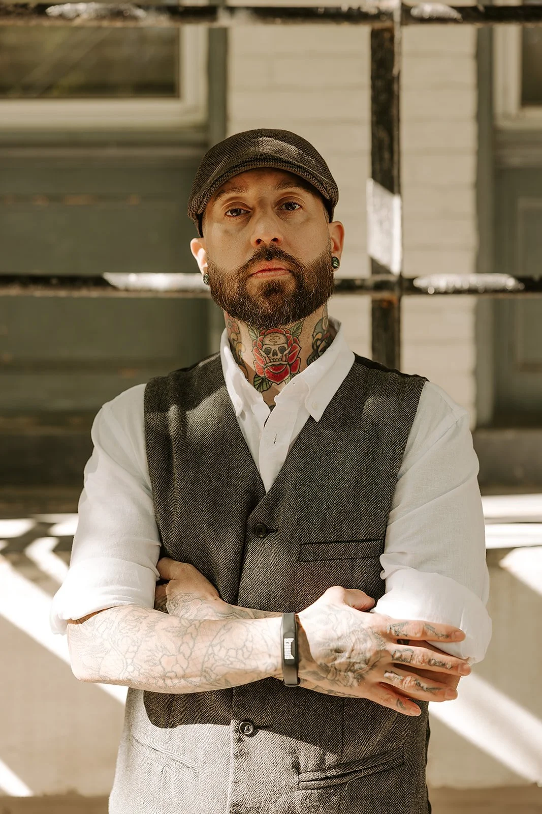 A man with a beard and tattoos on his arms, wearing a gray fedora, blue shirt, and gray vest, leaning against a brick wall with arms crossed.