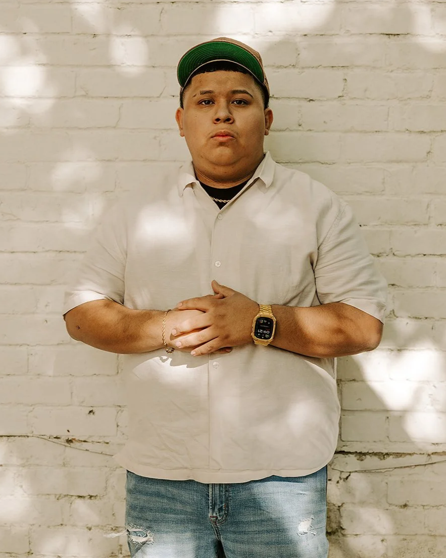 A young man standing with arms crossed against a brick wall, wearing a white cap and dark polo shirt.