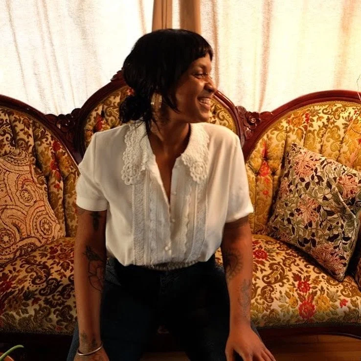 A Black Person with a Black mullet haircut sits on an antique floral couch.