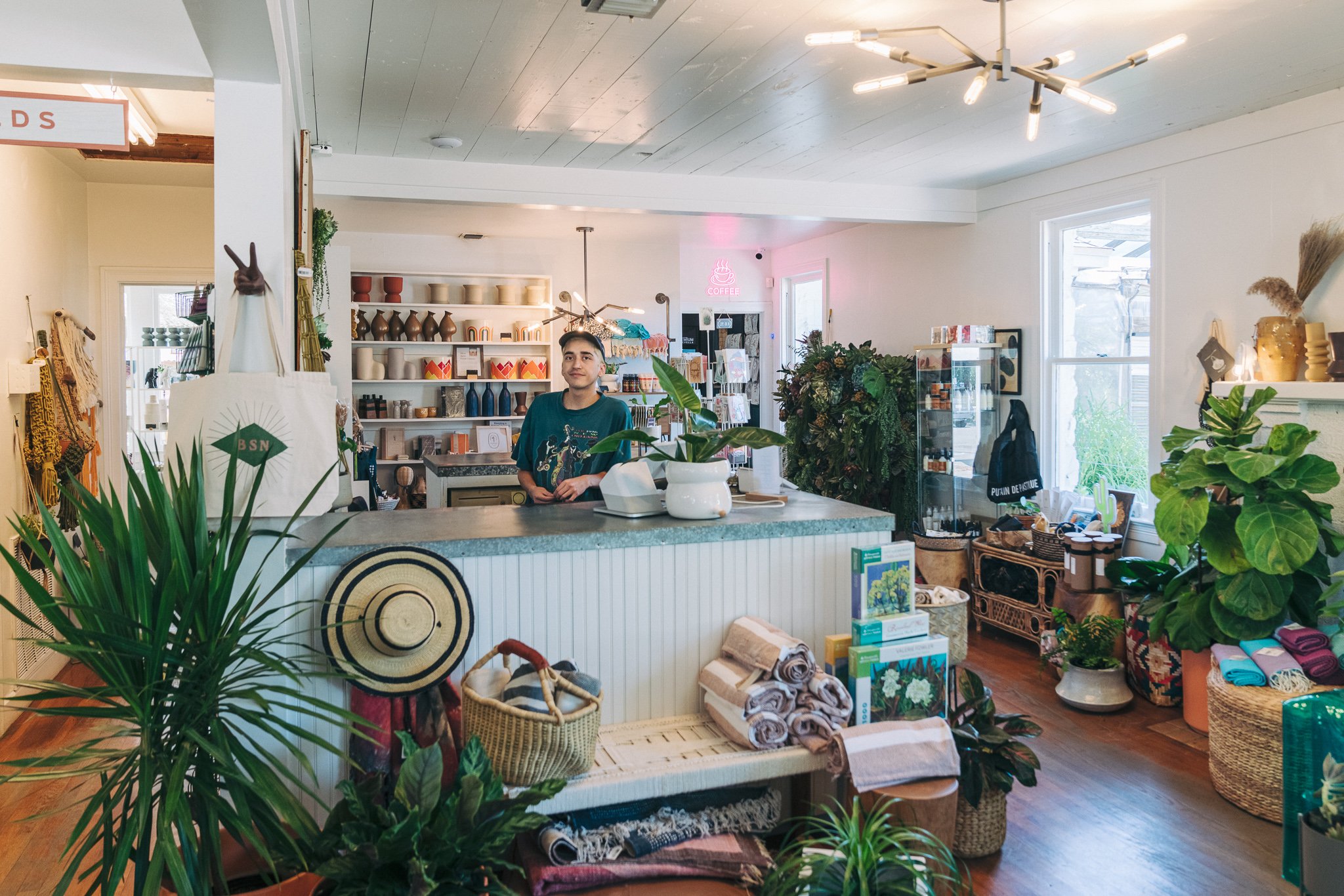 Interior of a cozy store with plants, shelves of pottery, textiles, and a counter with a person standing behind it.