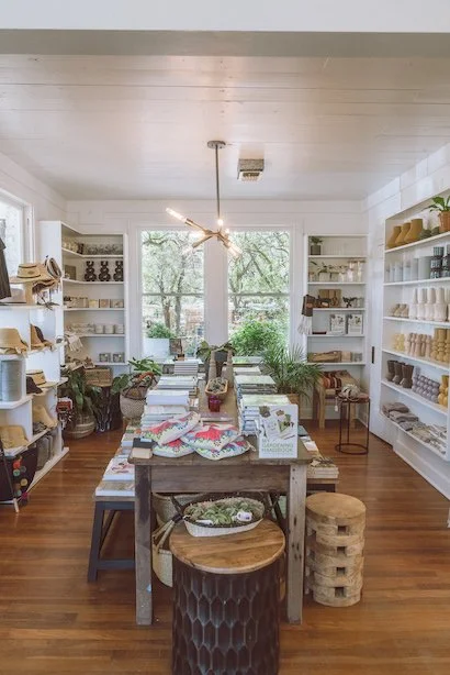 Charming boutique store with rustic wood furniture, plants, and various home decor items on display, featuring shelves with baskets, candles, and textiles under a modern pendant light.