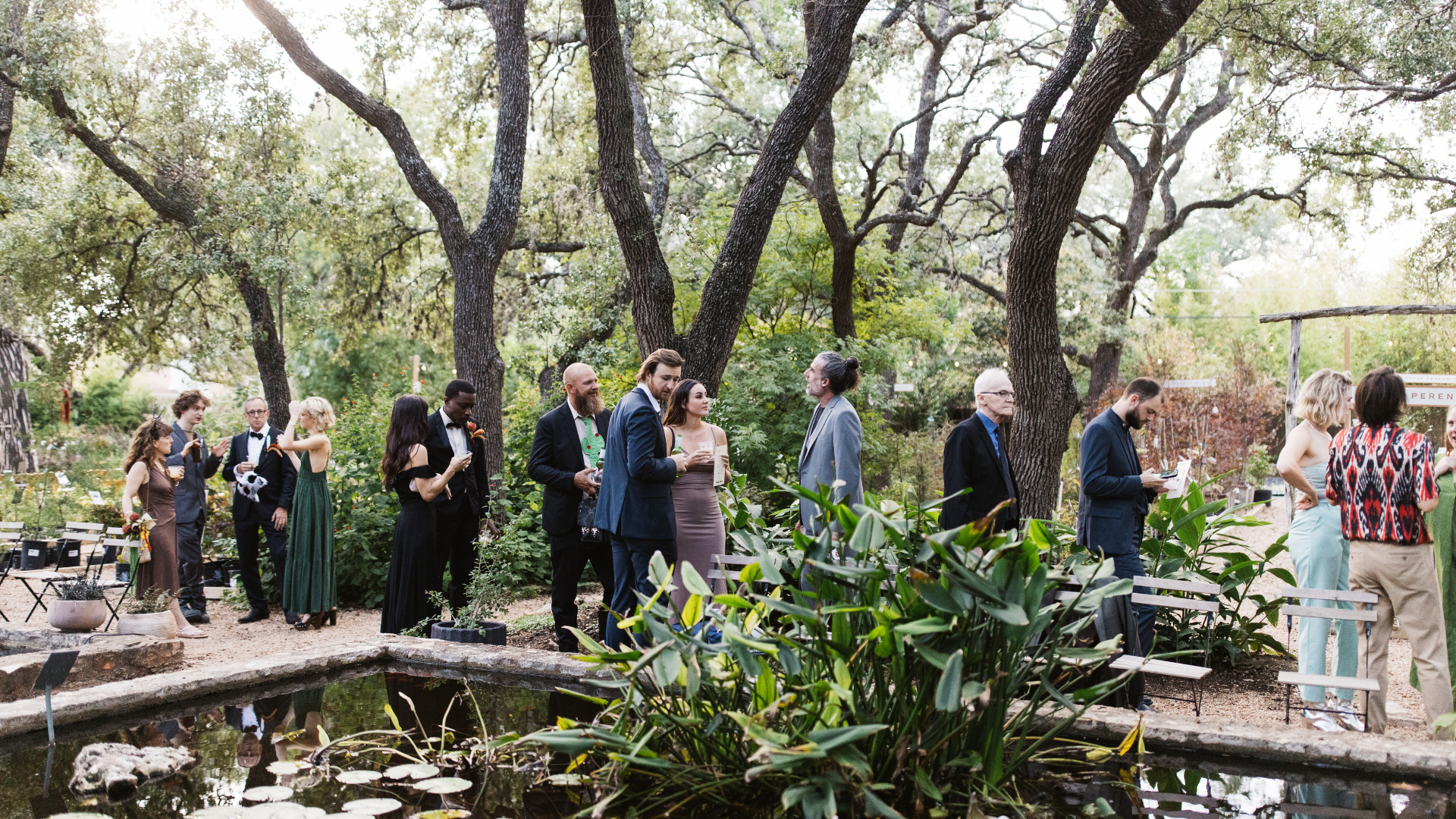 Group of people at an outdoor event in a garden setting, wearing formal and semi-formal attire, socializing around a pond with trees in the background.