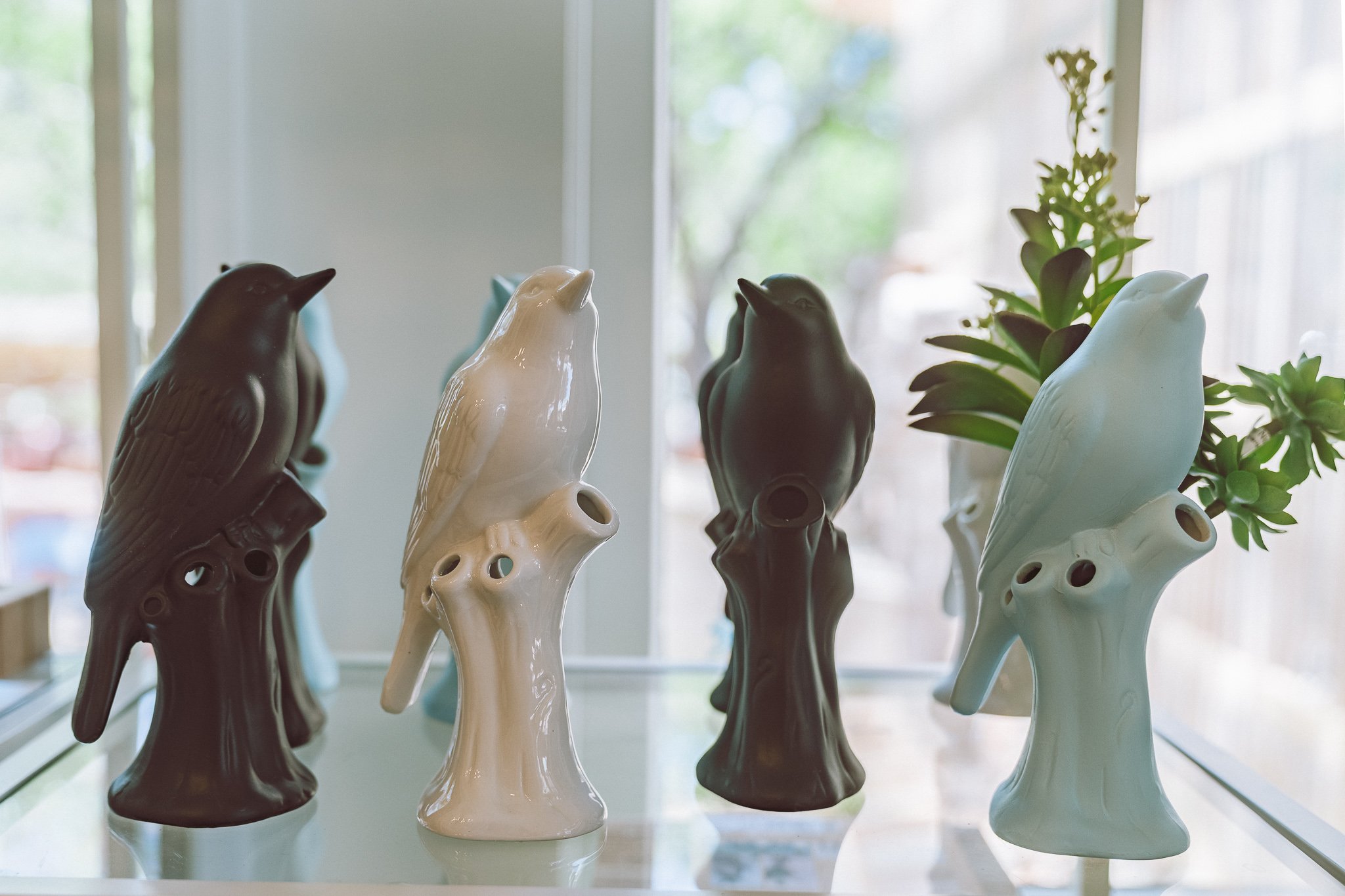 Ceramic bird figurines on tree branch stands with small plants on a window shelf.