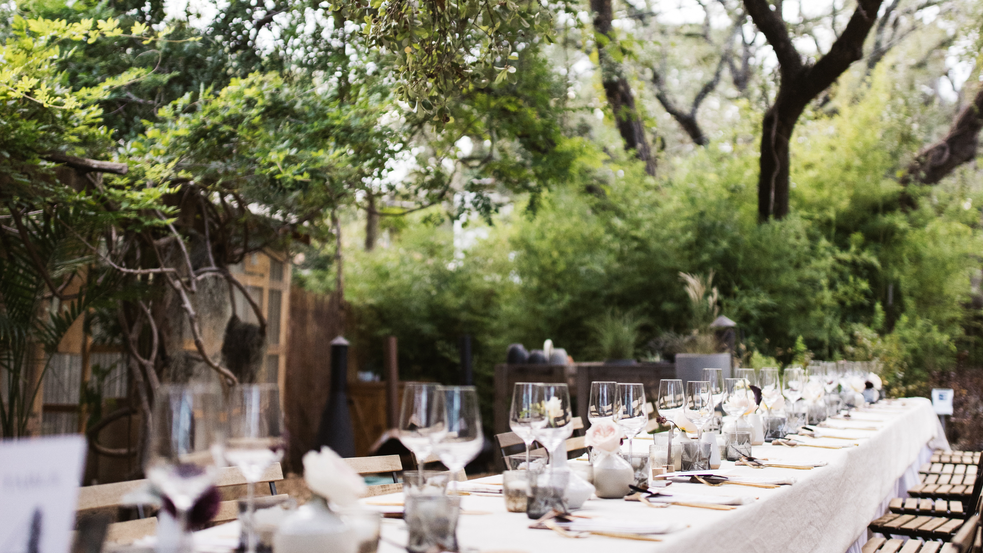 Outdoor dining table set for an event with white tablecloth, glasses, and floral arrangements, surrounded by trees and greenery.
