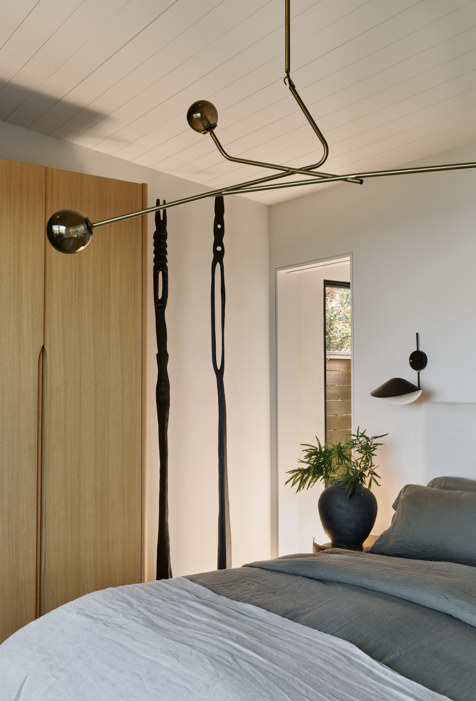 Modern bedroom with a bed, potted plant, black wall-mounted lamp, and unique ceiling light fixture with two black spheres and metal rods.