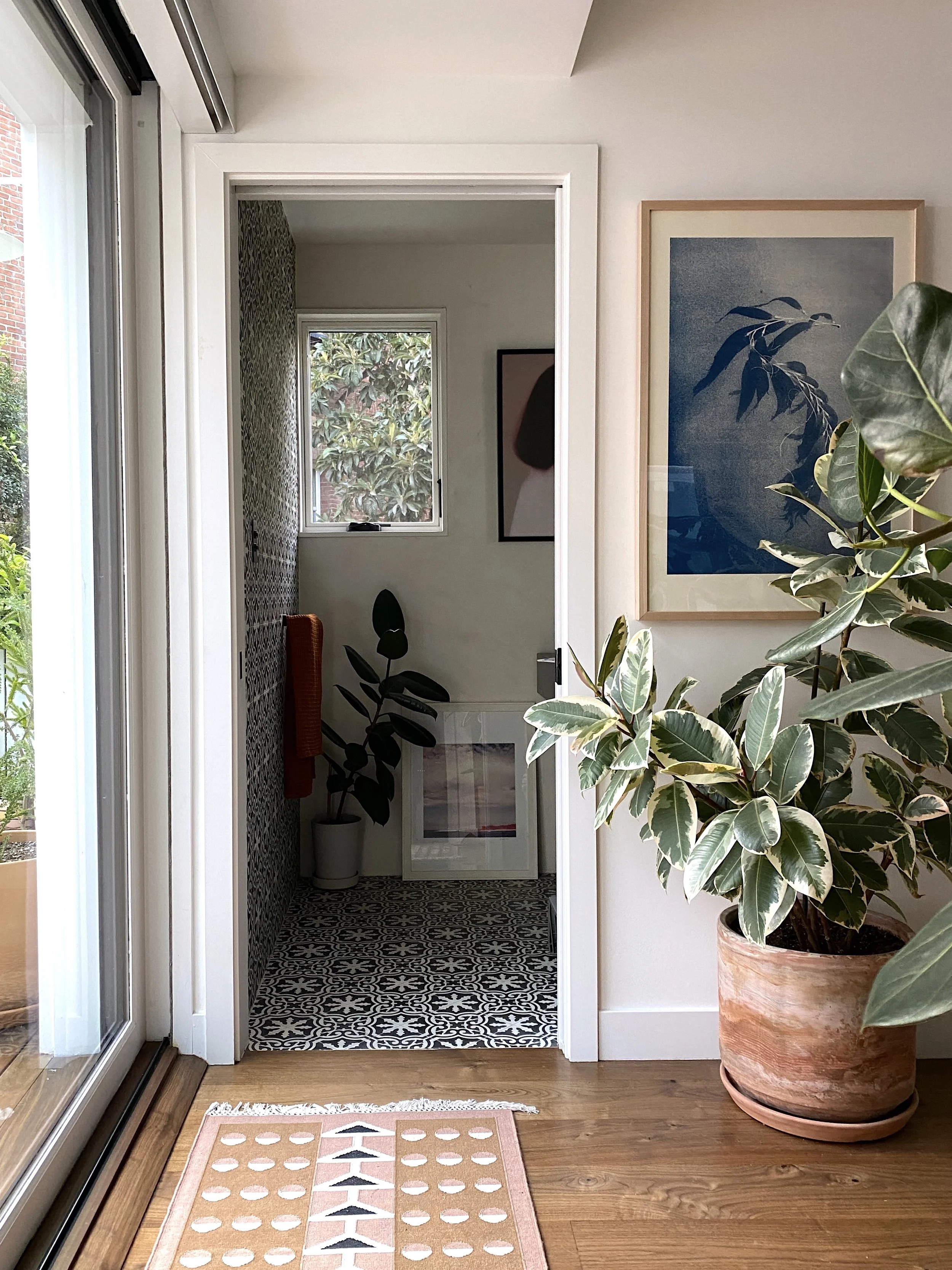 Los Angeles interior Design. Interior view of a home featuring a wooden floor, a large potted plant, and artwork on the wall, with a view into a small room with patterned black-and-white floor tiles and plants