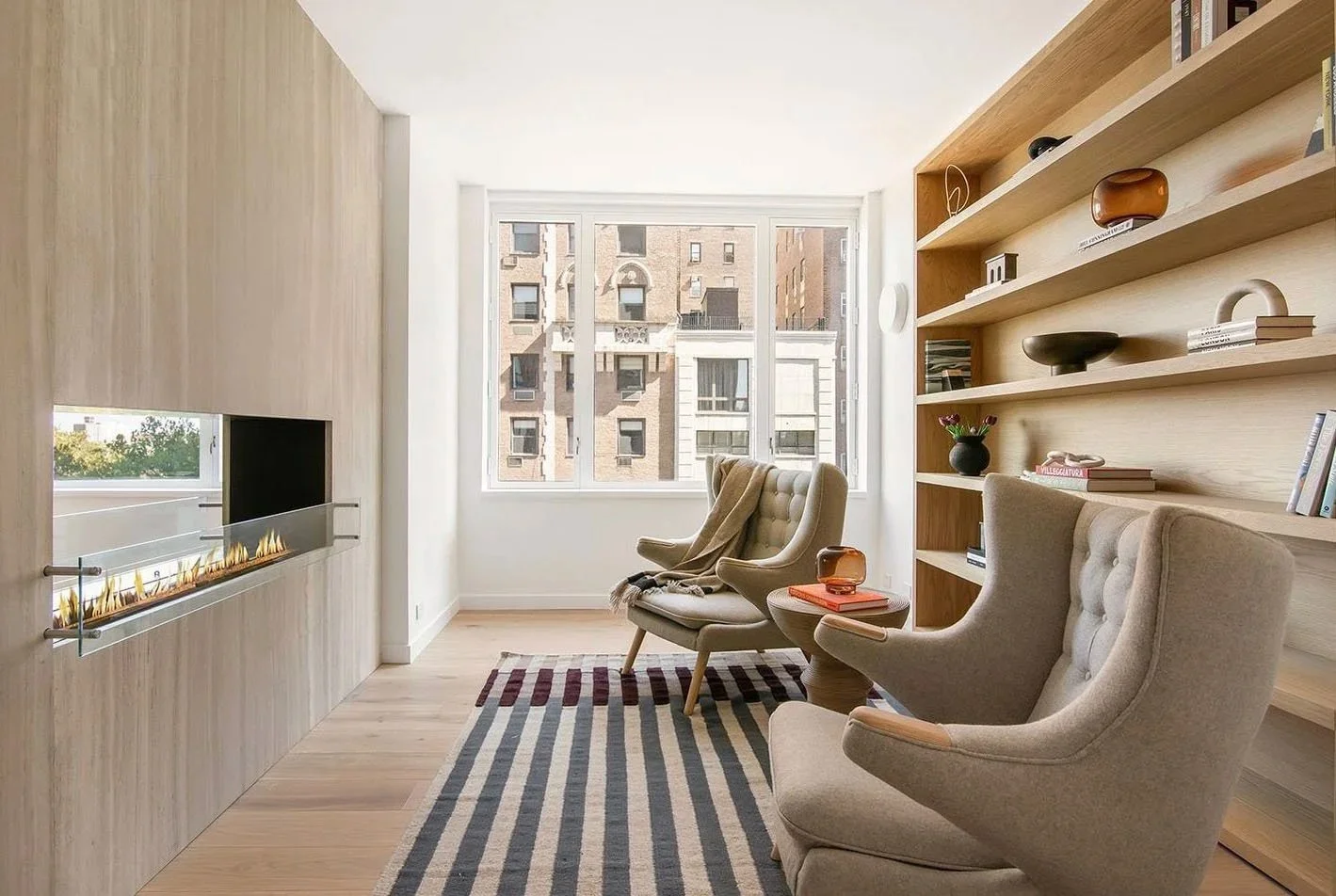 Bright living room with beige armchairs, a striped rug, a large window, a wooden bookshelf with decor, and a built-in fireplace.