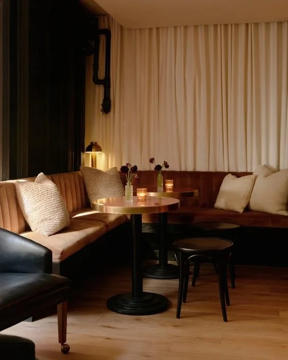 Cozy corner with a brown curved sofa, beige pillows, a small round table with two vases of flowers, and a black chair, in a softly lit room with wood flooring and cream curtains.