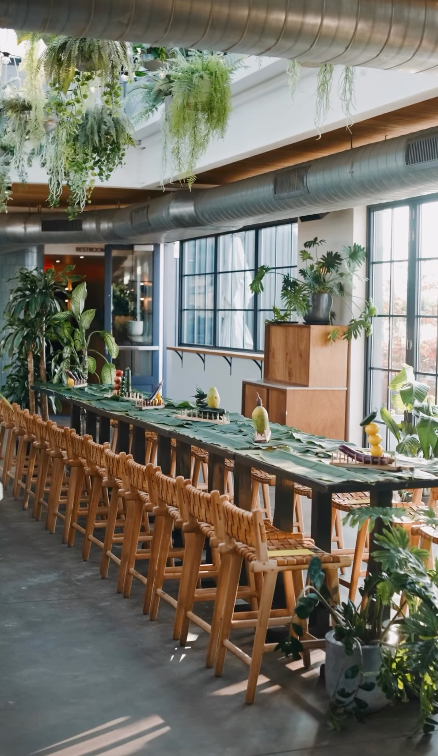 Rockaway Hotel Restaurant Design. A long dining table in a bright, modern restaurant decorated with leafy plants and tropical motifs, surrounded by wooden chairs.