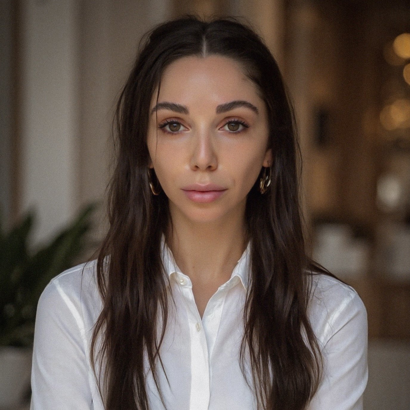 A portrait of a young woman with long dark hair, wearing a white button-up shirt and gold hoop earrings, looking directly at the camera with a neutral expression, in an indoor setting with blurred warm lighting and decor in the background.