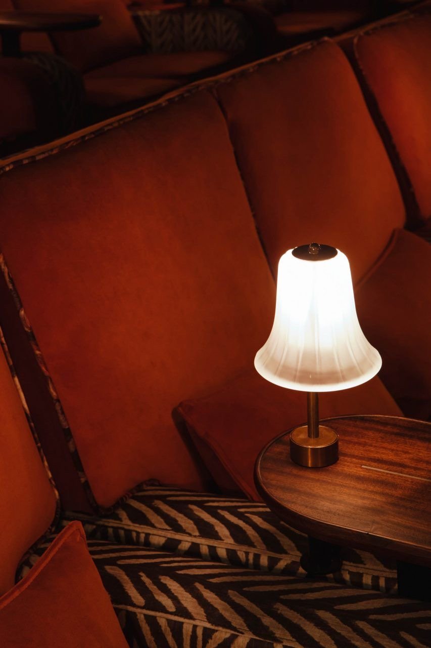 A lit desk lamp with a white lampshade on a round wooden table, against a background of orange upholstered cushions and a zebra print rug.
