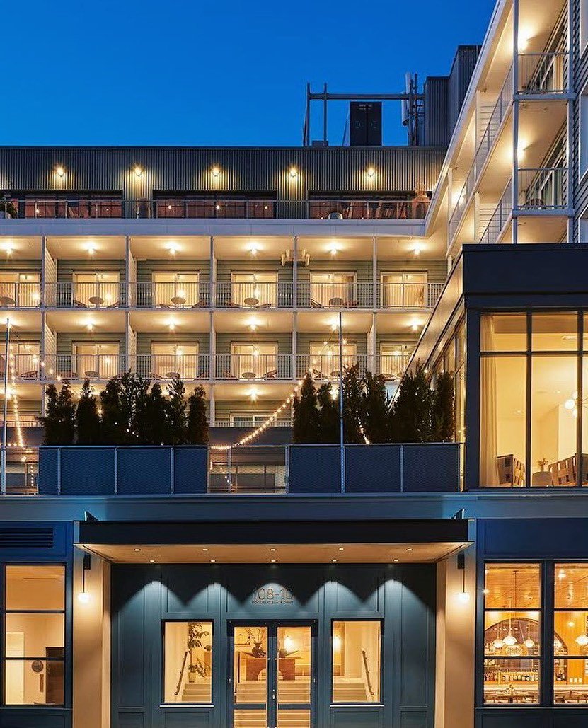 A multi-story modern hotel building at night, with illuminated balconies and large glass windows, featuring outdoor string lights and indoor seating visible through the windows.