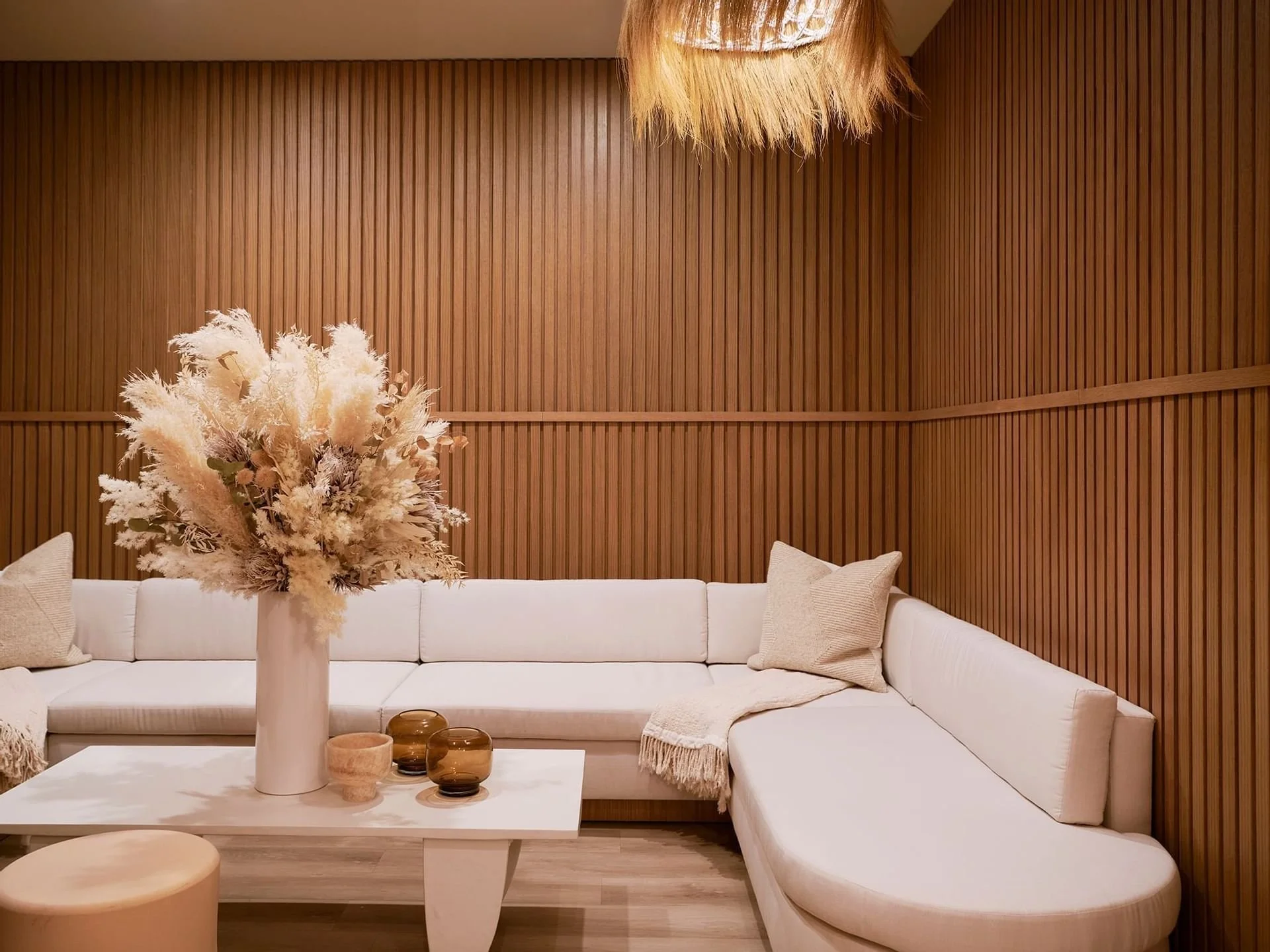 A modern living room with a white sectional sofa, beige pillows, a white coffee table with a large white vase with dried flowers, and a wooden paneled wall.